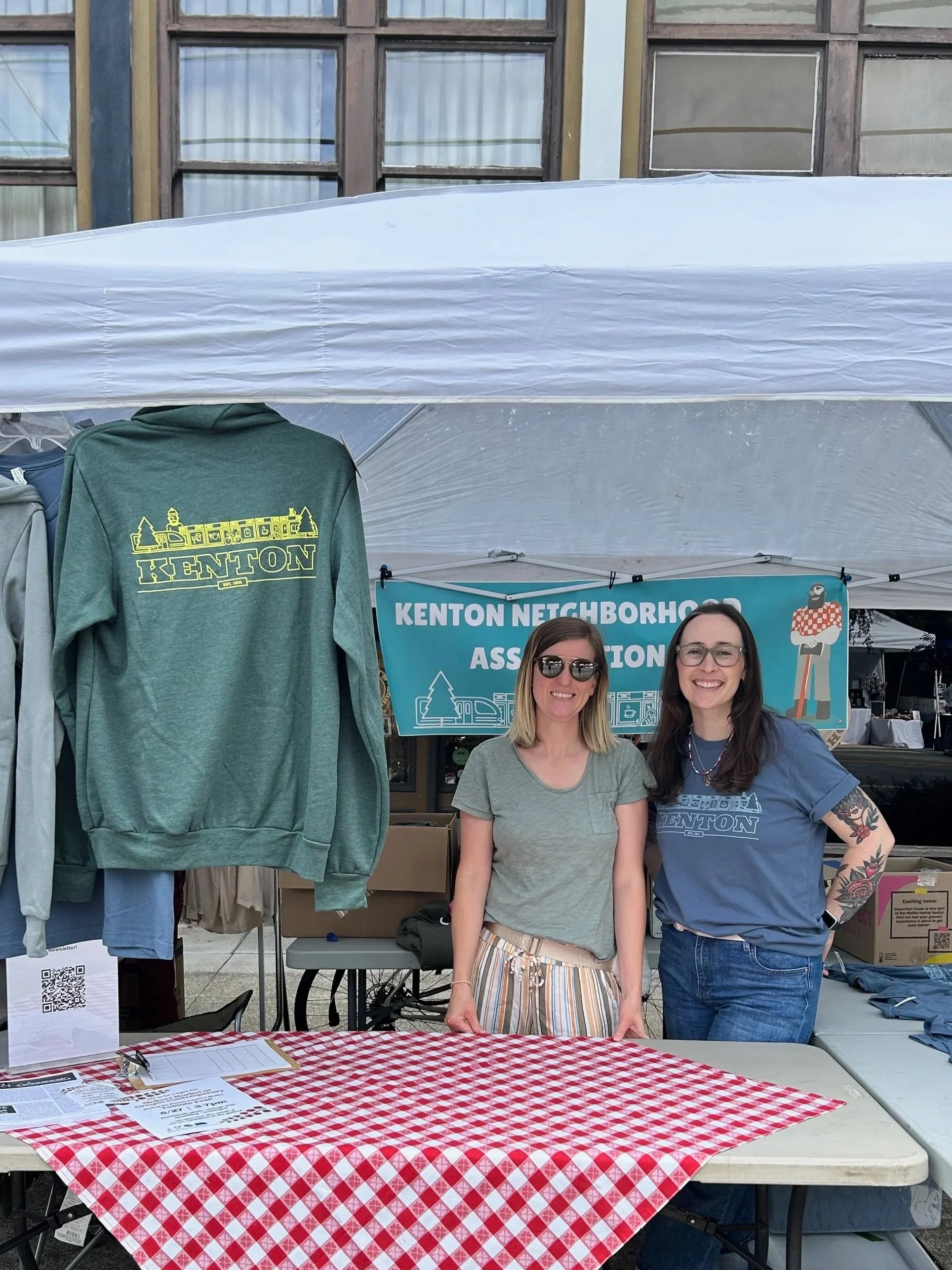 Two women smile while staffing a Kenton Neighborhood Association booth with merch on display.