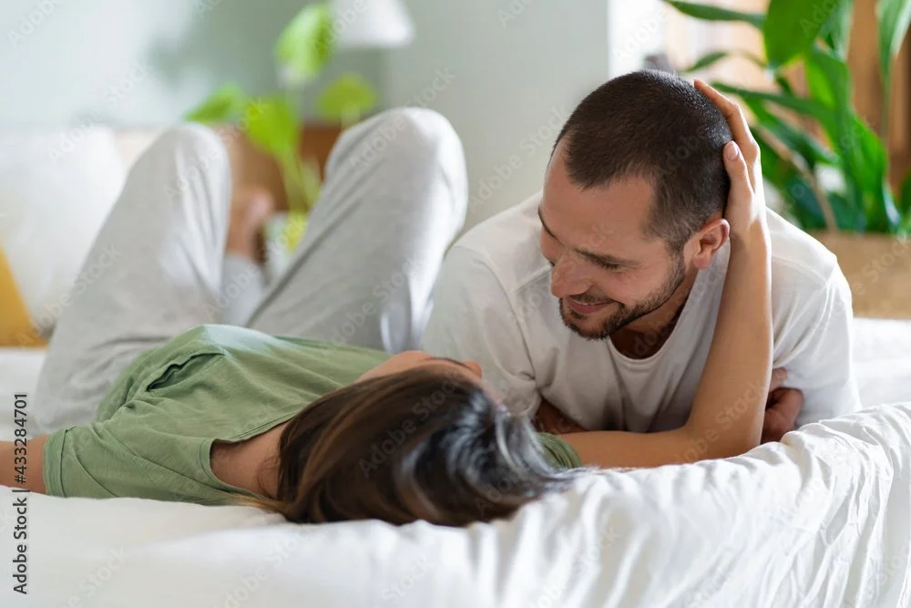 A man and woman lying on a bed, smiling and looking at each other. The man is holding the woman's head with one hand, and they appear to be having a joyful moment.