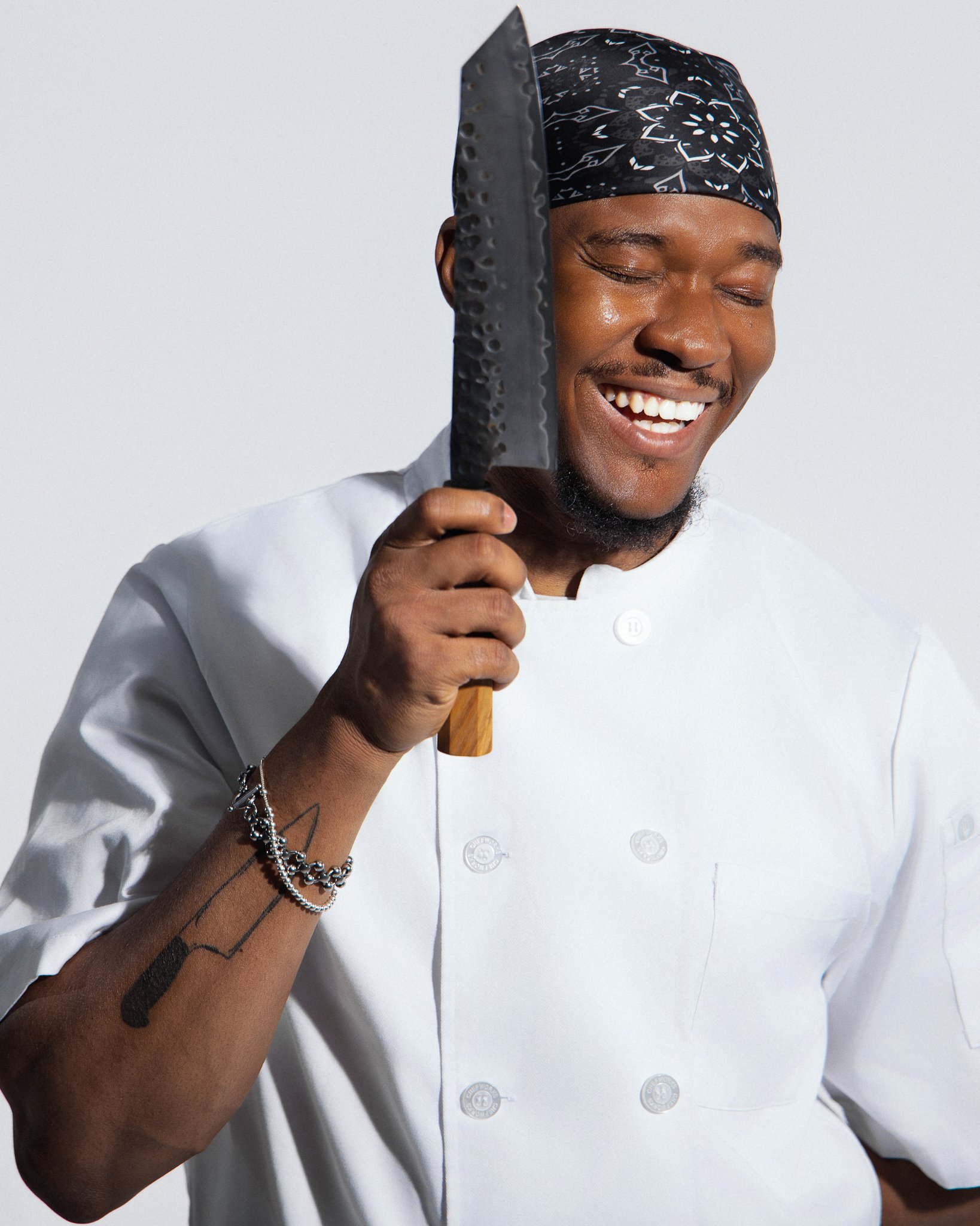 A chef smiling and holding a large kitchen knife close to his face, wearing a bandana and a white chef's coat.