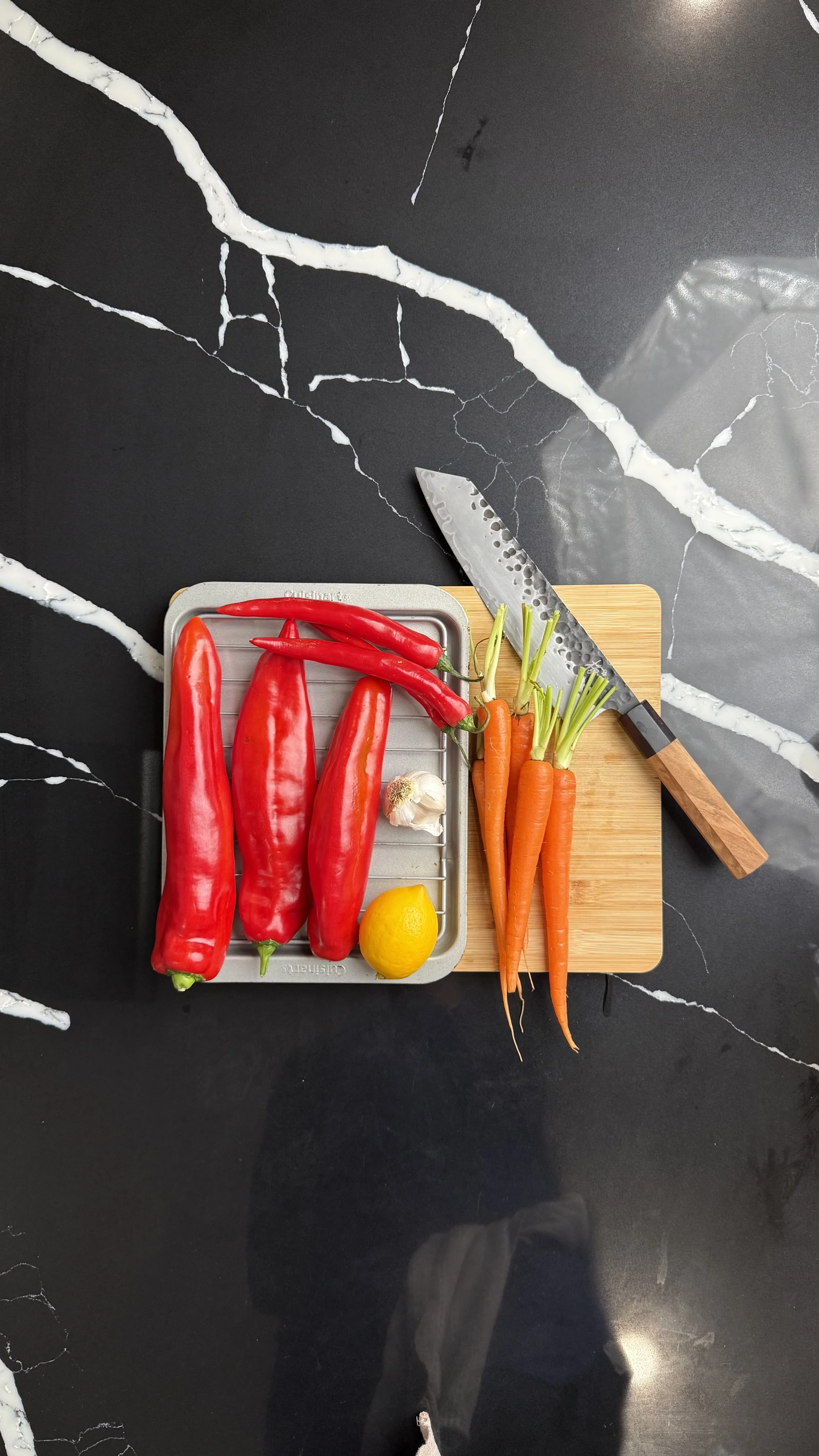 Red peppers, red chilies, carrots, garlic, lemon, and chef knife arranged on cutting board and tray for mojo rojo and roasted carrots.