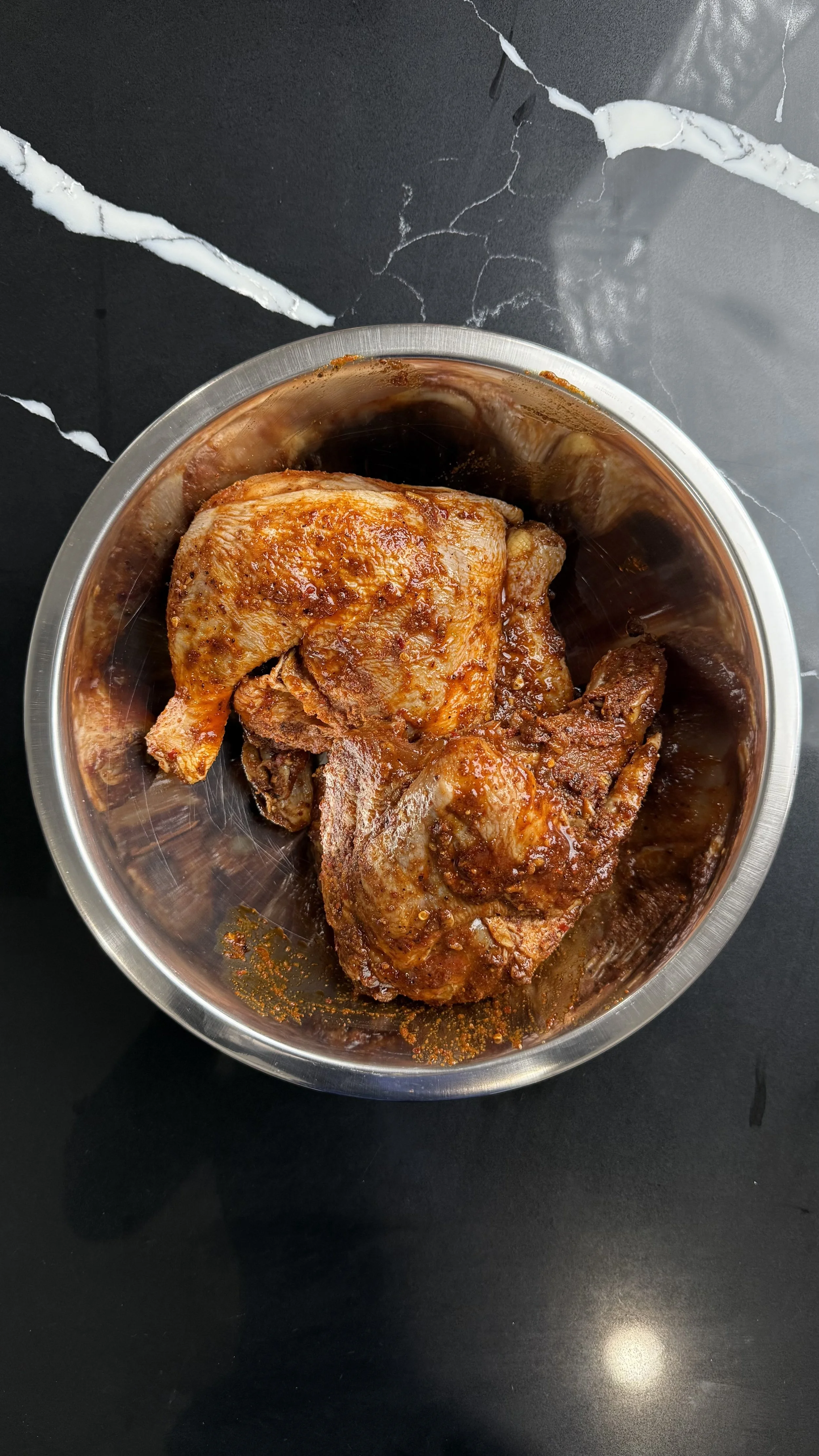 Half chicken coated in spice rub resting in a stainless steel bowl before air frying.