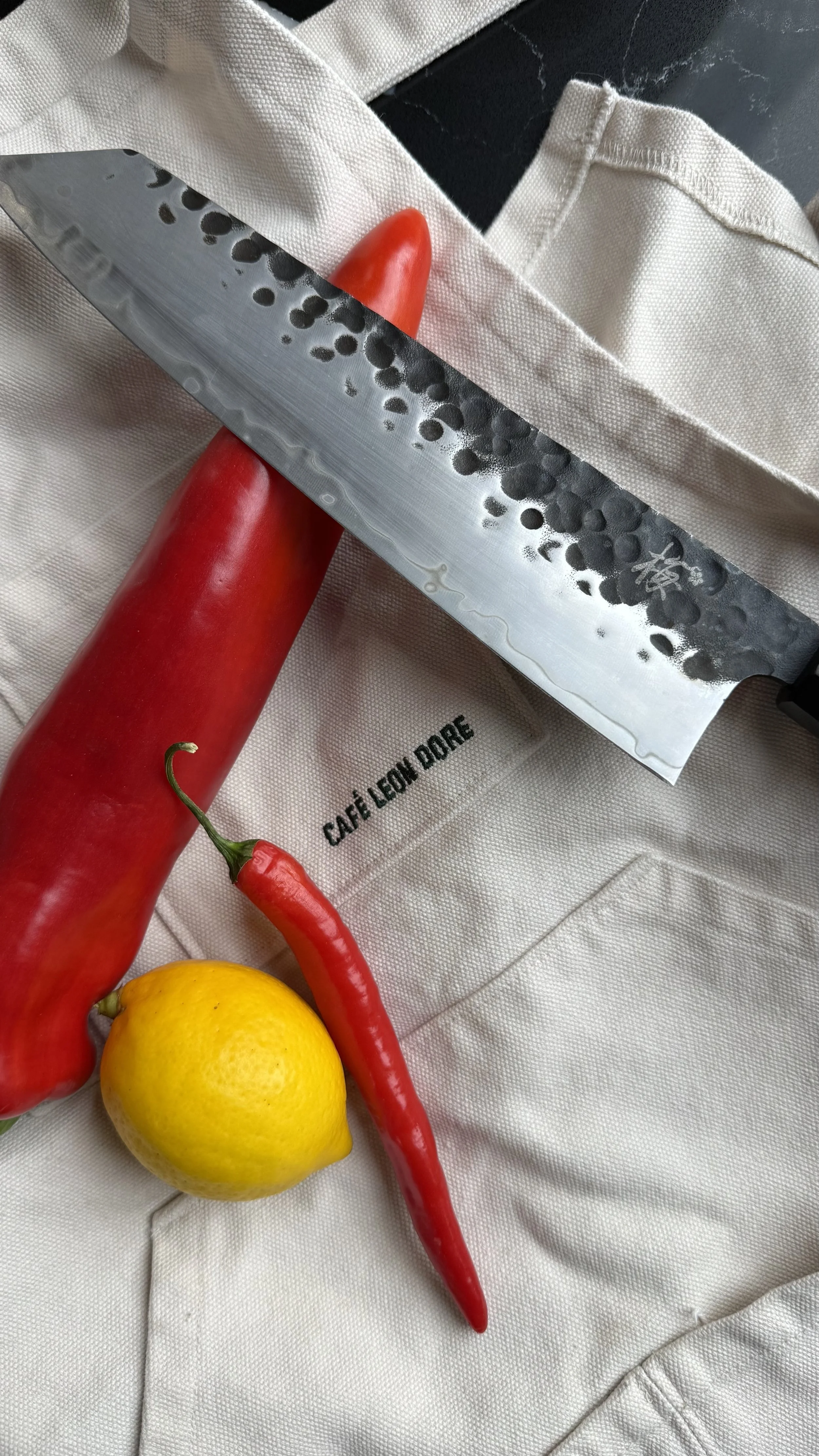 Fresh red Mediterranean peppers, red chili, and lemon styled beside a Japanese chef knife on a canvas apron.