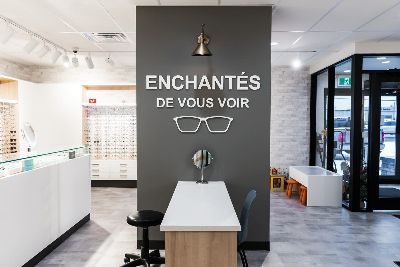 Interior of an eyewear store with a sign that reads 'Enchantés de vous voir' and a pair of glasses icon on a gray accent wall, with tables, chairs, and display shelves for glasses.