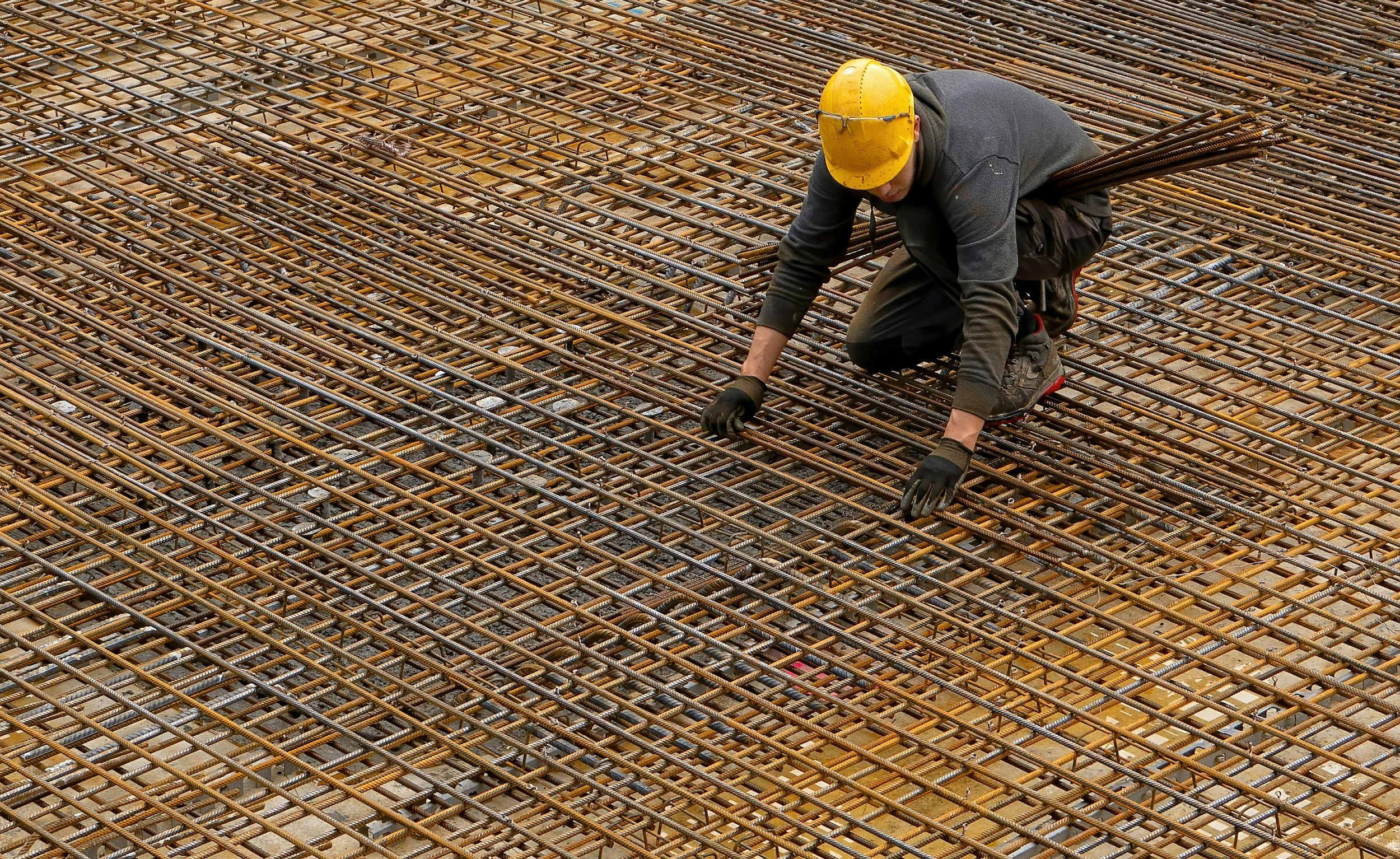 A construction worker wearing a yellow safety helmet, black gloves, and dark work clothes kneels on a large steel rebar grid at a construction site.