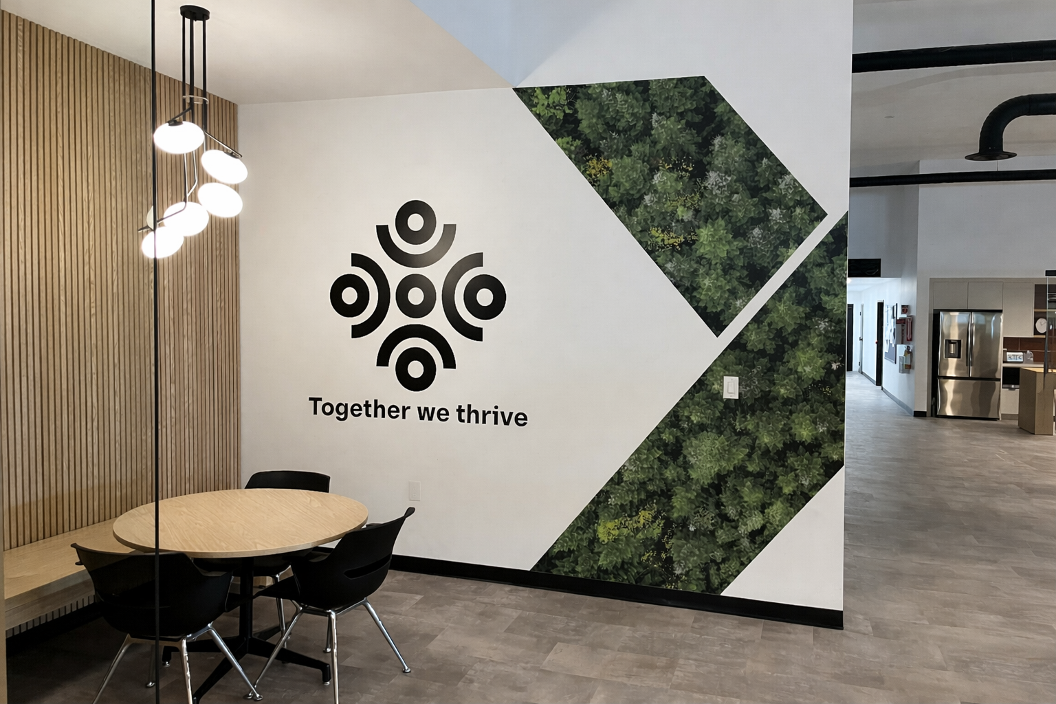 Office interior with wooden wall, modern lighting fixture, round table with three black chairs, mural with green foliage, and the phrase 'Together we thrive' on the wall.