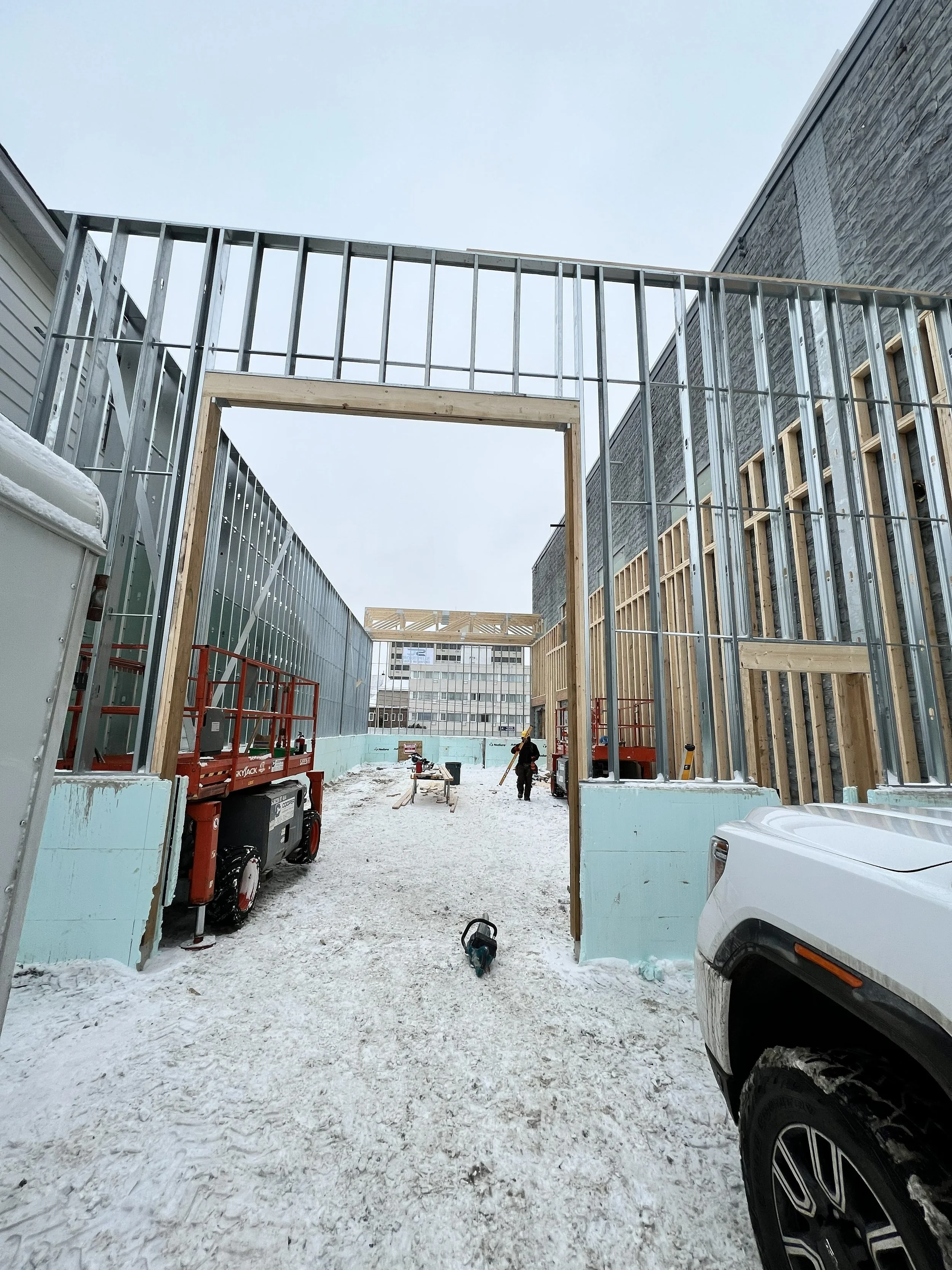 Construction site with a partially built structure, steel framing, wooden supports, construction workers, construction equipment, and snow-covered ground.
