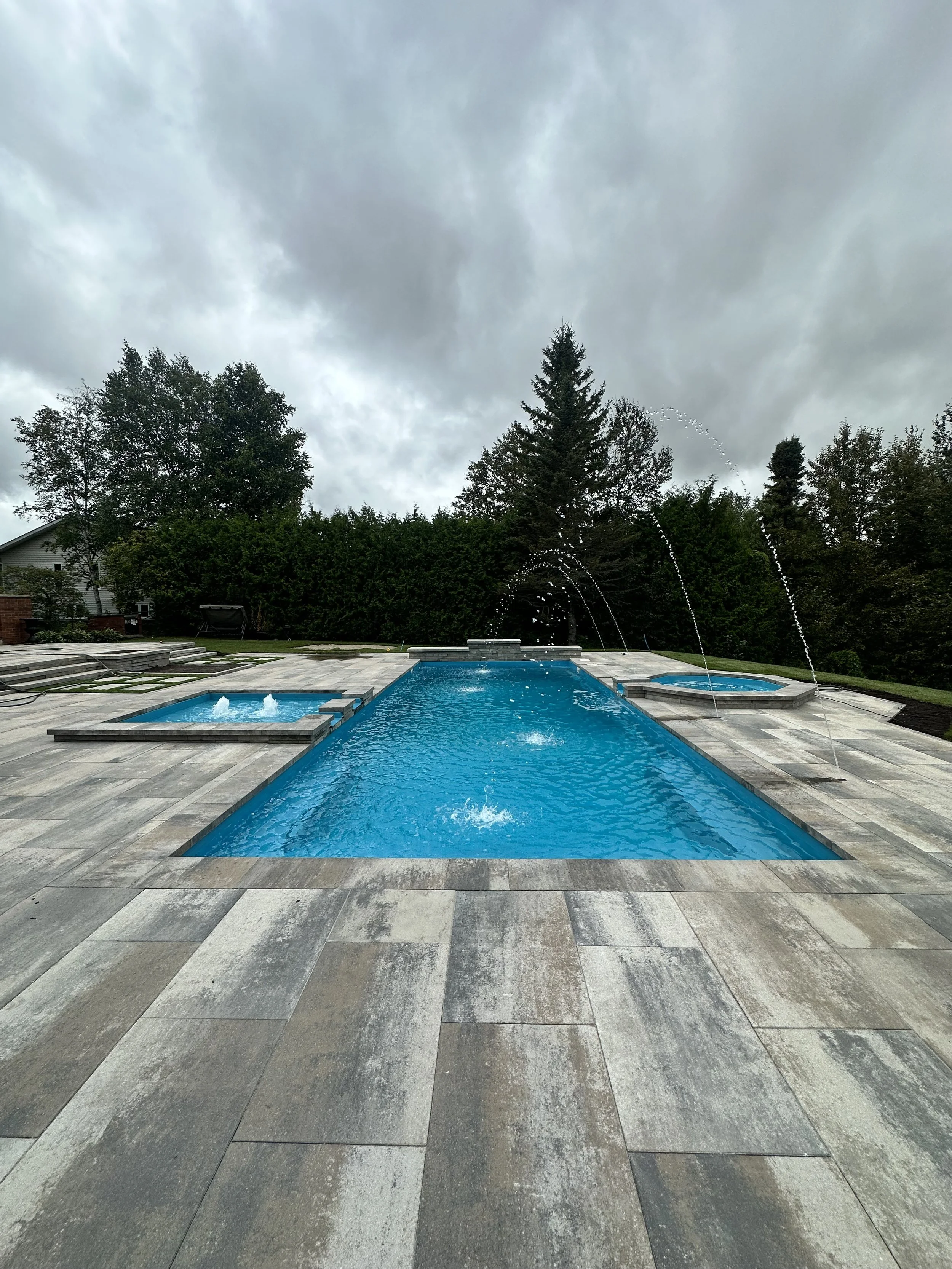 An outdoor swimming pool with a hot tub and fountain jets in a backyard surrounded by trees under a cloudy sky.