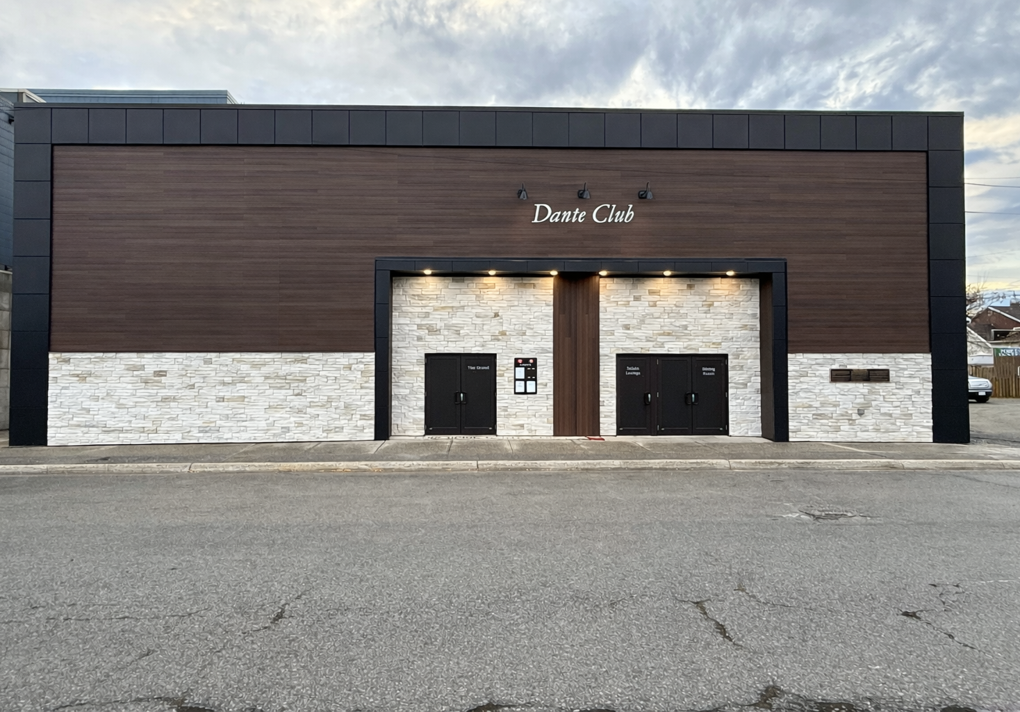 Front view of a modern building with a sign that reads 'Dante Club,' featuring a facade with wood and stone finishes, three black doors, and external lighting on a cloudy day.