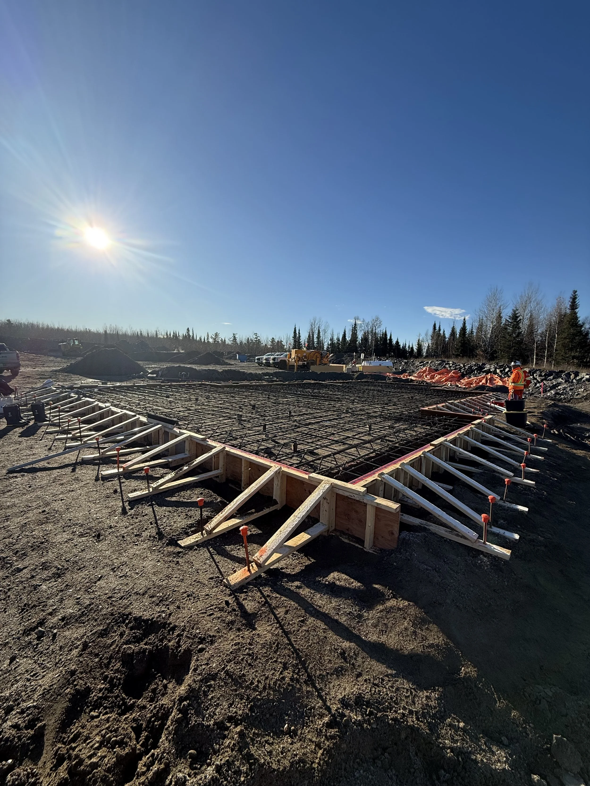 Construction site with a concrete foundation being prepared, setting rebar and wooden formwork, under a clear blue sky with the sun shining brightly.
