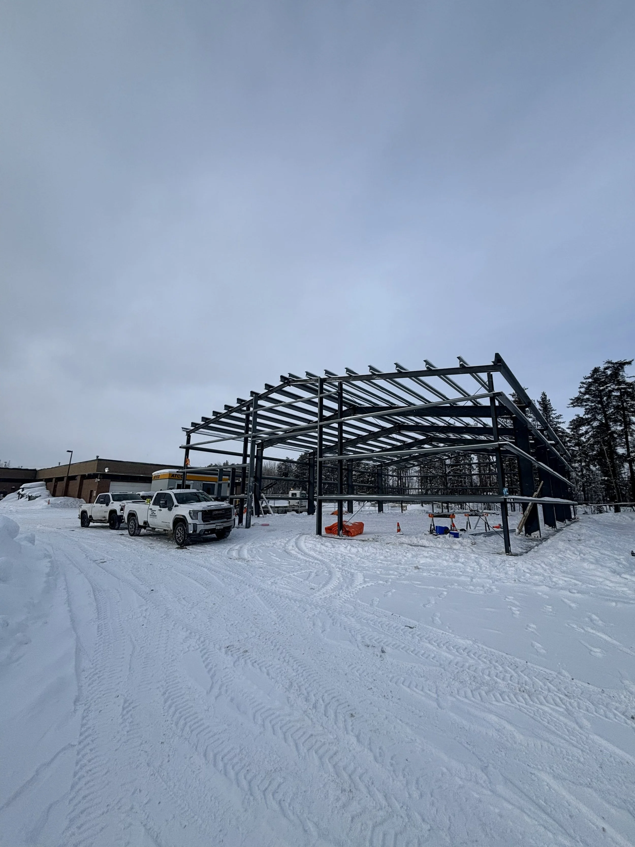 Construction site with a steel frame structure in a snow-covered area, several white trucks parked nearby, and trees in the background.