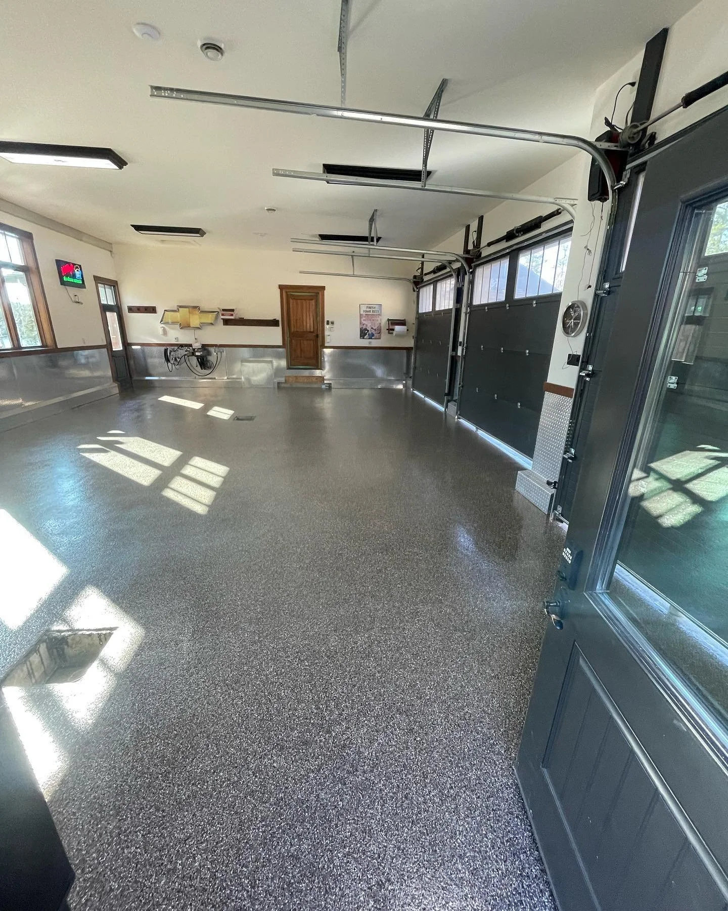 An empty modern garage with polished epoxy floors, two large garage doors, a wooden door, windows letting in natural light, and minimal wall decorations.
