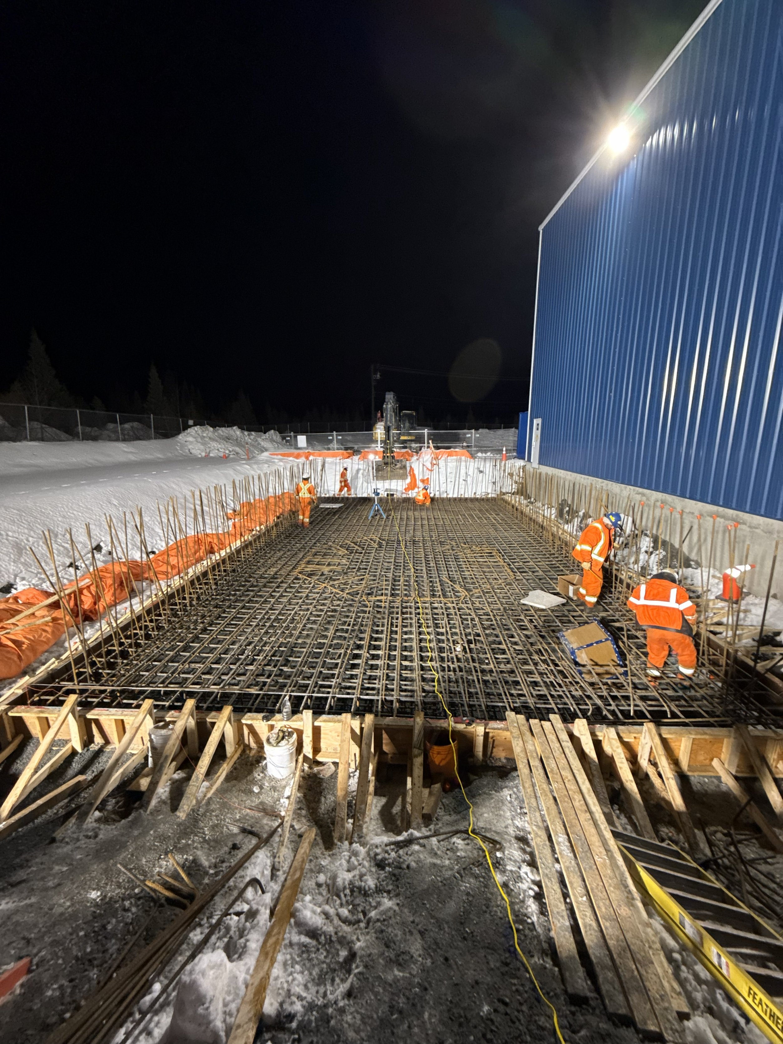 Construction workers in orange safety gear working on a steel rebar framework at night, with snow on the ground and a large blue building to the right, illuminated by bright outdoor lights.