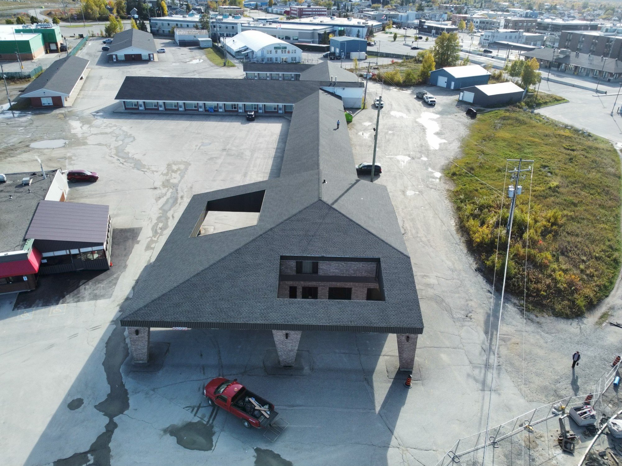 An aerial view of a commercial building with a large parking lot, surrounded by other commercial buildings and greenery. The building has a distinctive L-shaped roof with large open cutouts.