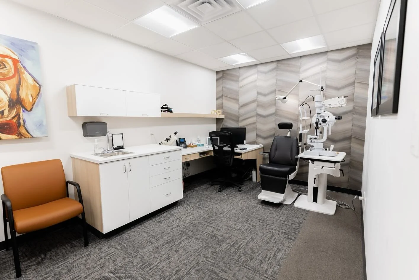 Medical examination room with ophthalmology equipment, a desk with a computer, a black office chair, a brown waiting chair, and artwork on the wall.