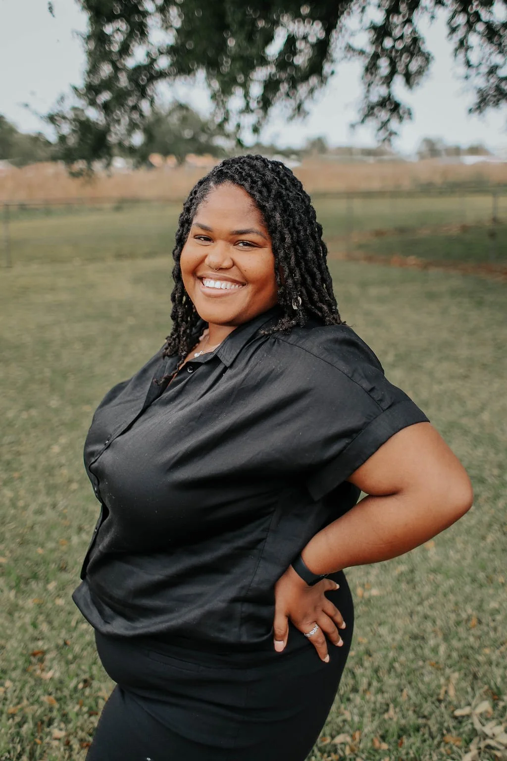 A woman with dark, curly hair smiling outdoors with trees and a fence in the background. She is wearing a black shirt and has her hand on her hip.