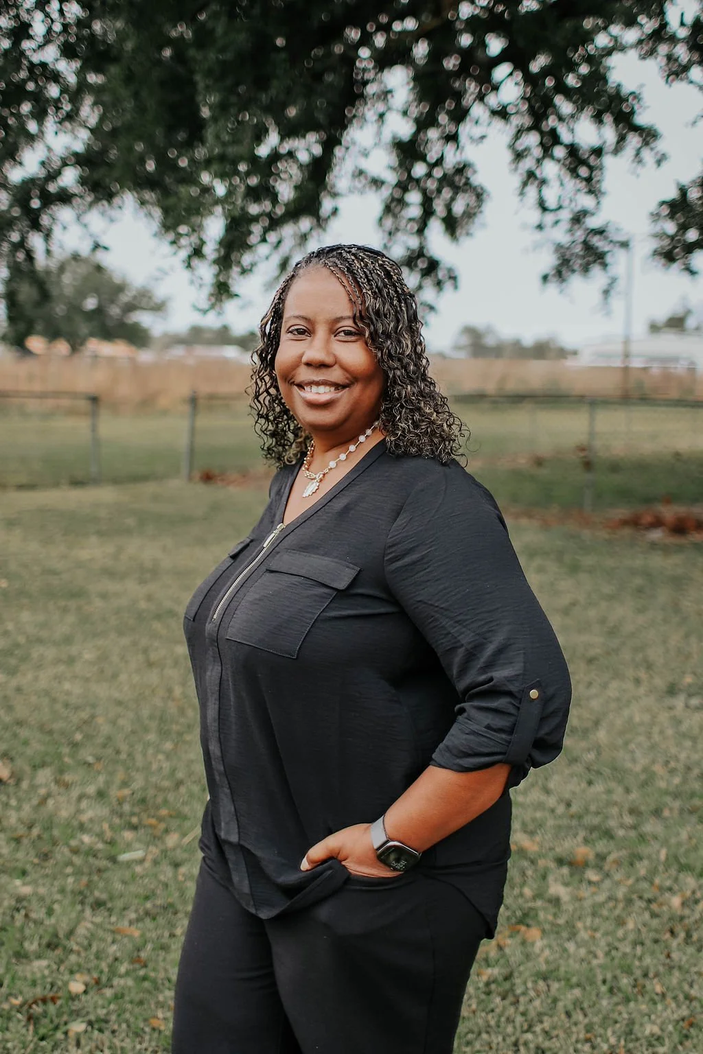 A woman smiling outdoors near a tree, wearing a black long-sleeve top, black pants, a watch, a pearl necklace, and a necklace with a leaf pendant, with her hand in her pocket.