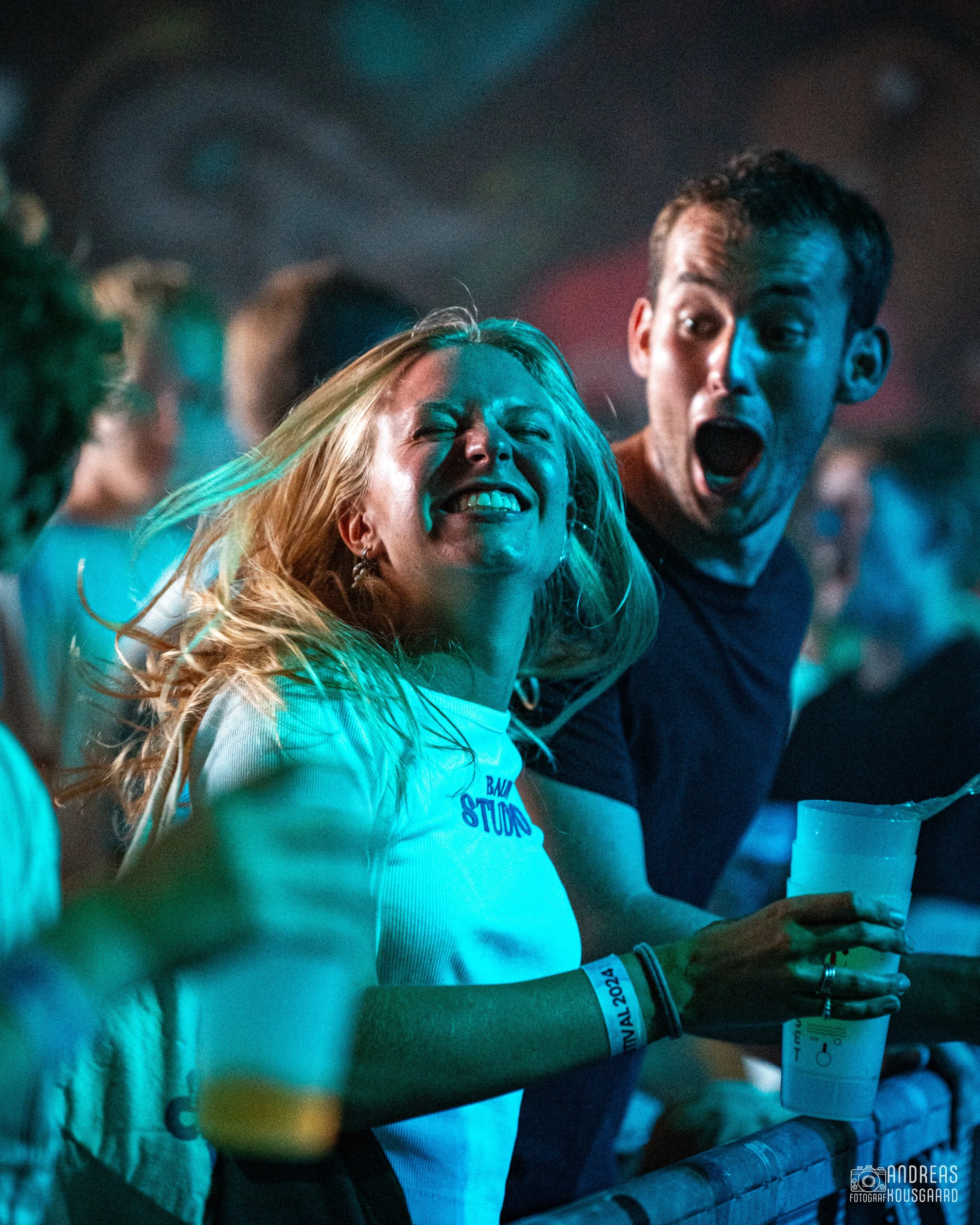 Two people dancing at a concert, a woman with blonde hair smiling and a man with short hair looking surprised, both holding drinks, with a colorful, blurred background.