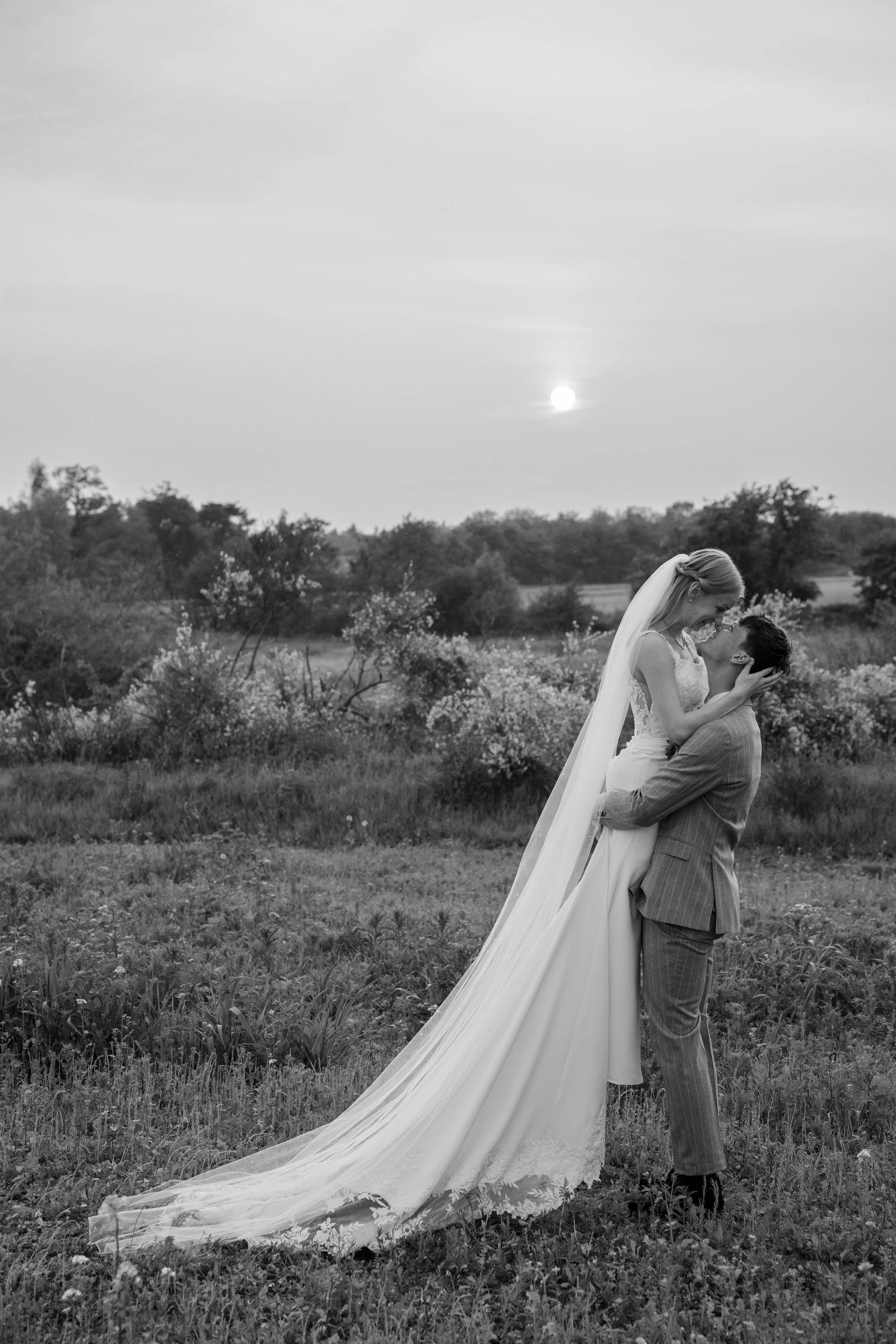 A bride and groom standing in a field during sunset, embracing and smiling at each other, with the bride wearing a wedding dress with a long veil and the groom in a pinstripe suit.
