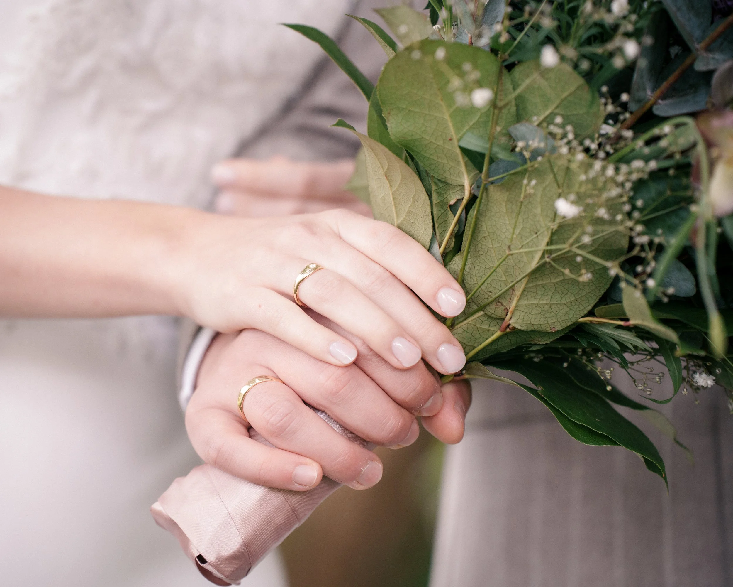 A couple's hands with wedding rings holding a bouquet of greenery and small white flowers.