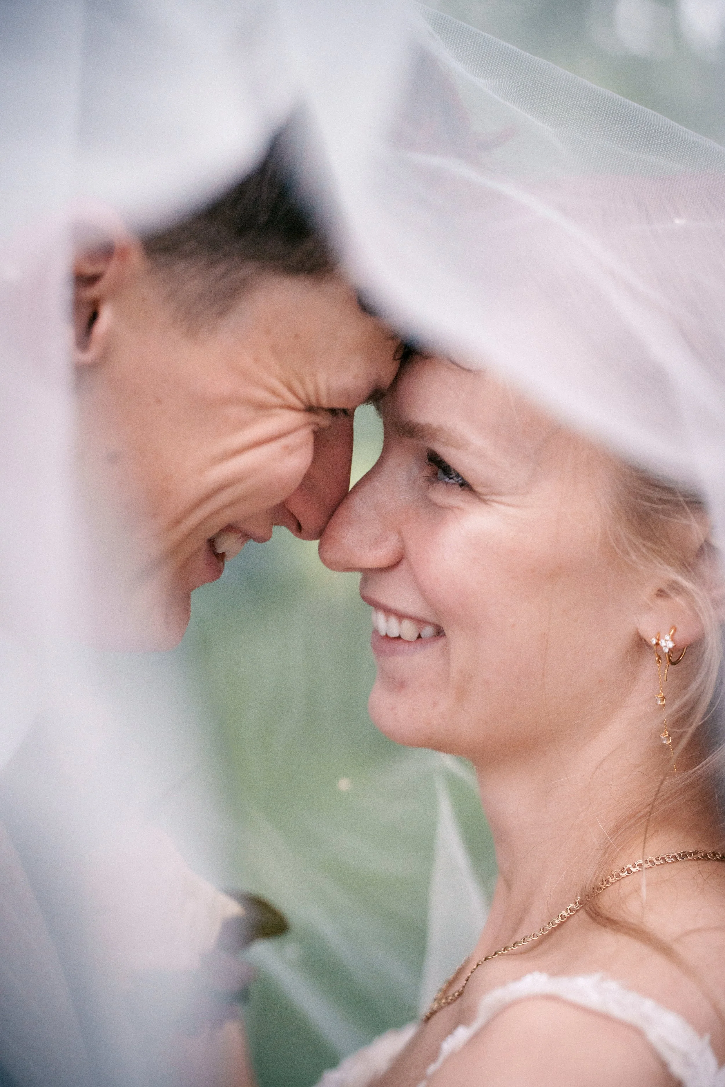 A close-up photo of a couple smiling and touching foreheads under a white veil during their wedding.