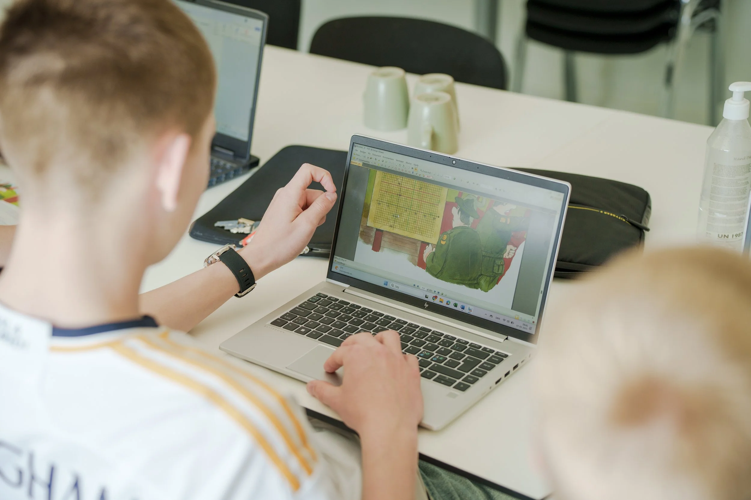 A person working on a silver laptop with a drawing of a person in a green uniform on the screen, sitting at a white table with two more laptops, a black bag, cups, a hand sanitizer bottle, and other items in a classroom or meeting room.