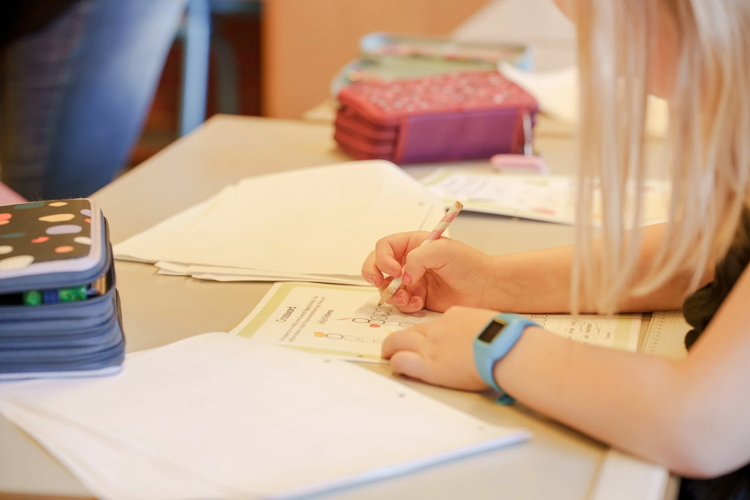 Child working on school worksheet with colored pencil, surrounded by books and notebooks on desk.