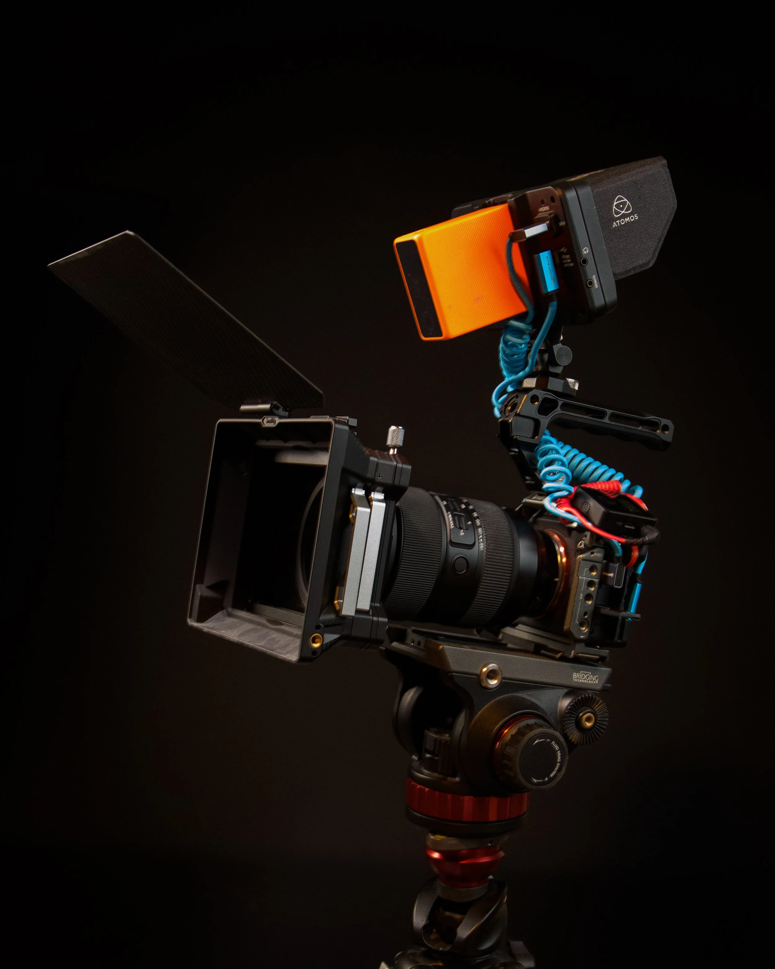 Camera mounted on a tripod with a matte box, attached monitor, and various cables, against a black background.