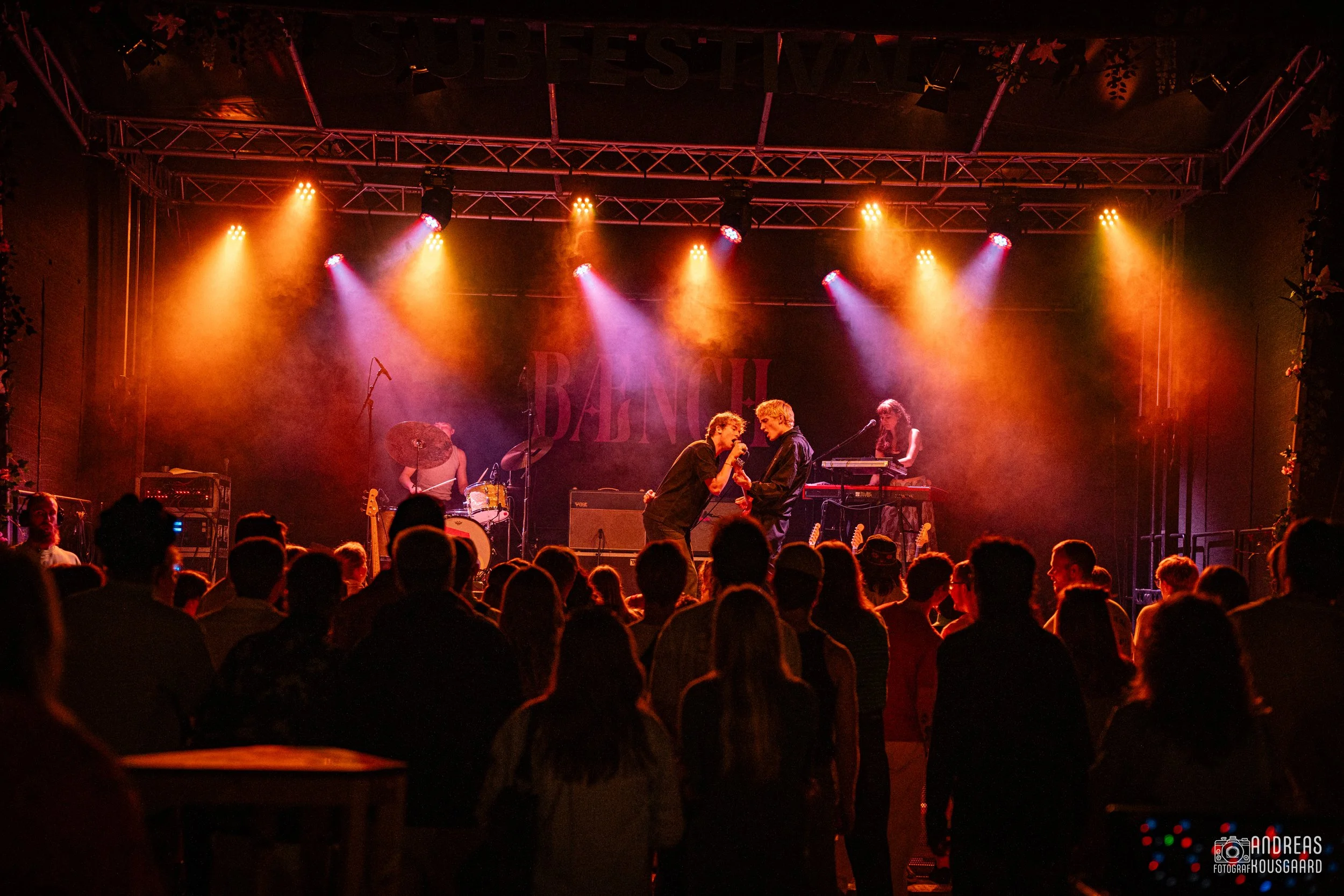 Live band performing on stage during a concert with colorful stage lighting and an audience watching.