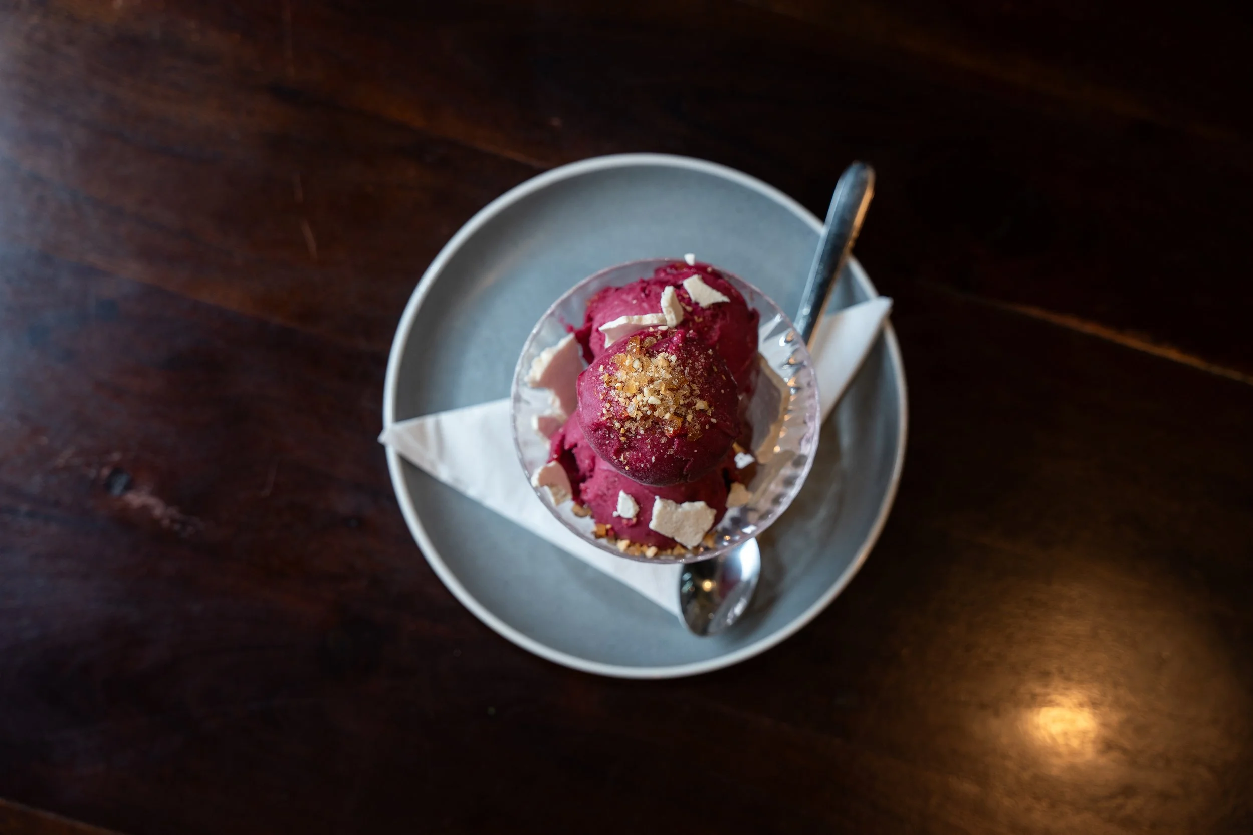 A dessert with two scoops of raspberry sorbet topped with chopped nuts in a glass cup, served on a gray plate with a spoon on a wooden table.