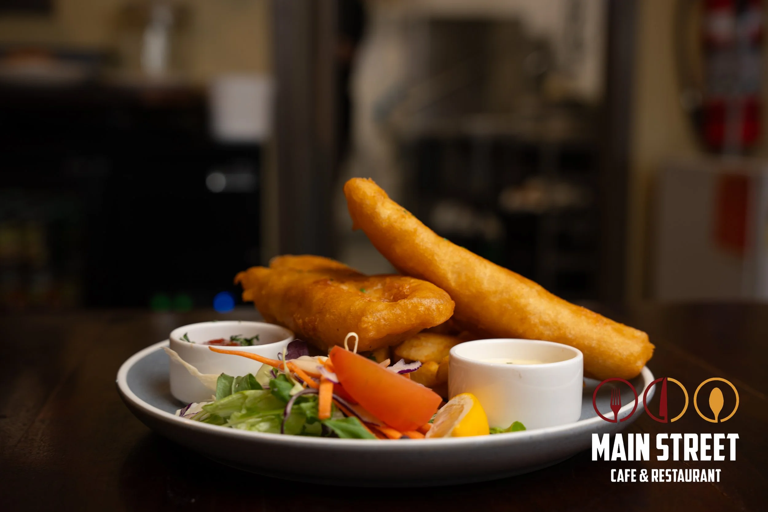 Fried fish and chips with salad, lemon wedge, and dipping sauces served on a white plate at Main Street Cafe & Restaurant.