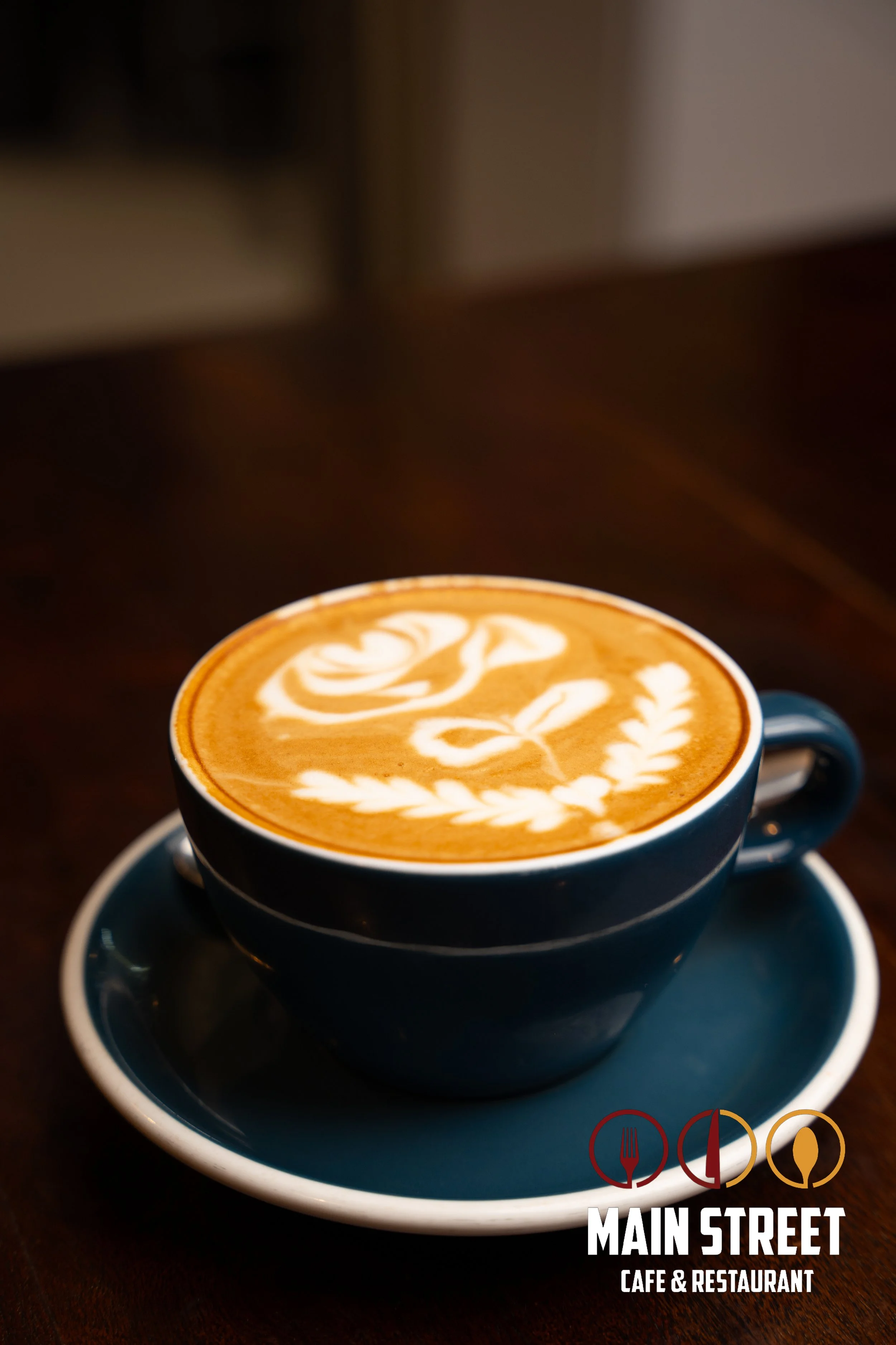 A cup of latte with latte art in a dark blue cup and saucer on a wooden table, with a logo for Main Street Cafe & Restaurant in the bottom right corner.