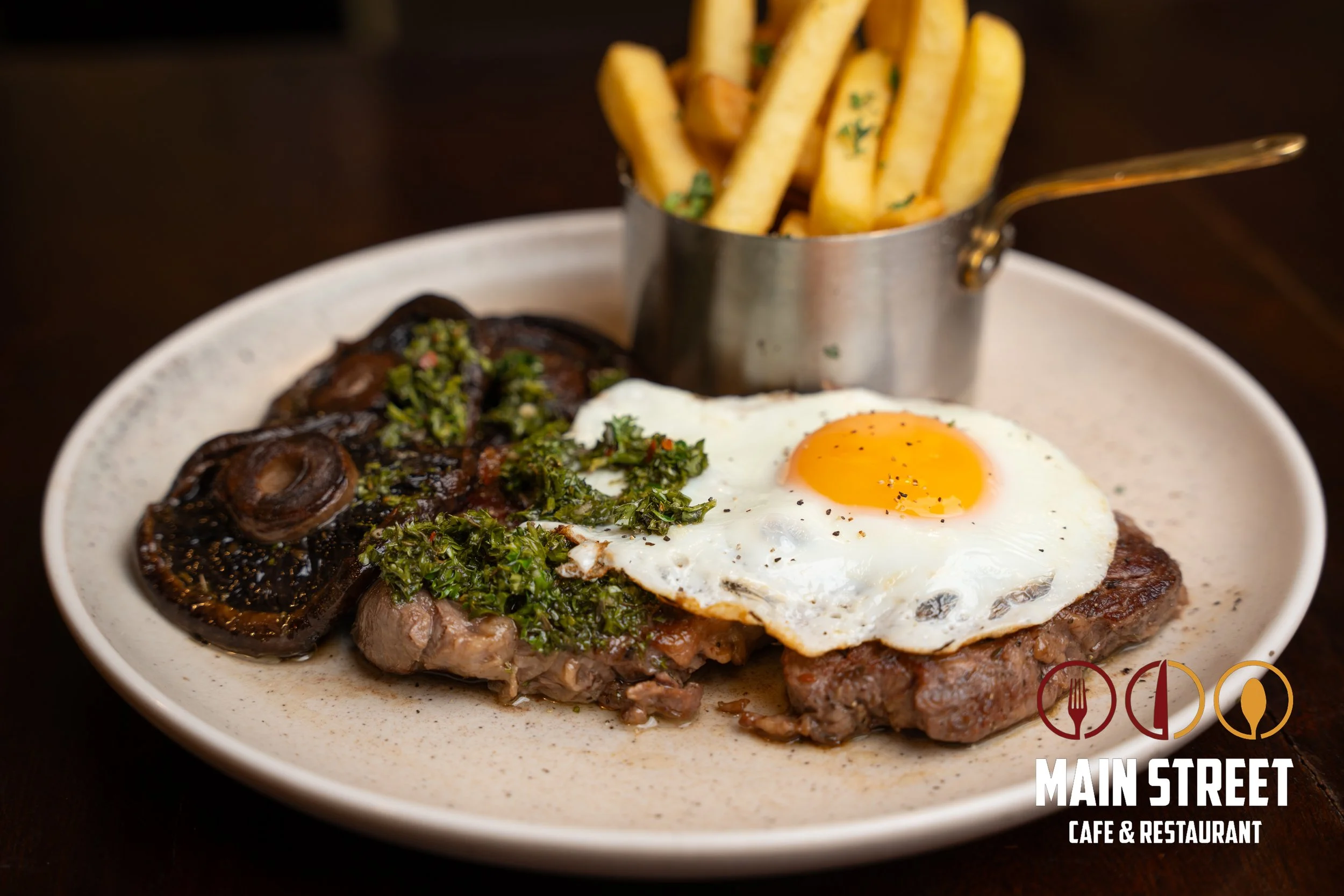 Steak dinner with fried egg on top, sautéed mushrooms, and grilled vegetables served with French fries in a metal cup on a white oval plate at Main Street Cafe & Restaurant