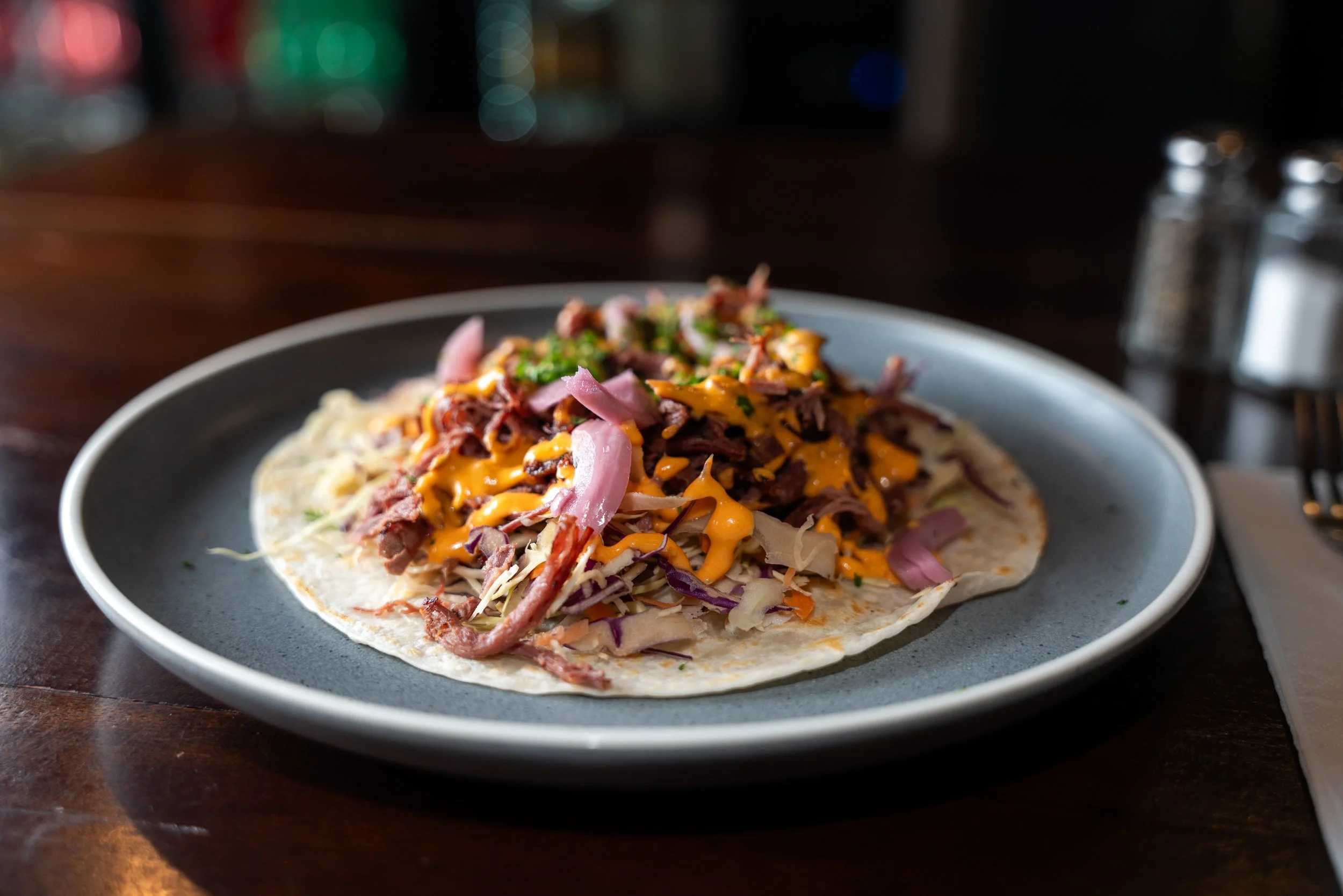 A taco with shredded meat, shredded cheese, chopped onions, and sauce on a soft tortilla on a dark plate.