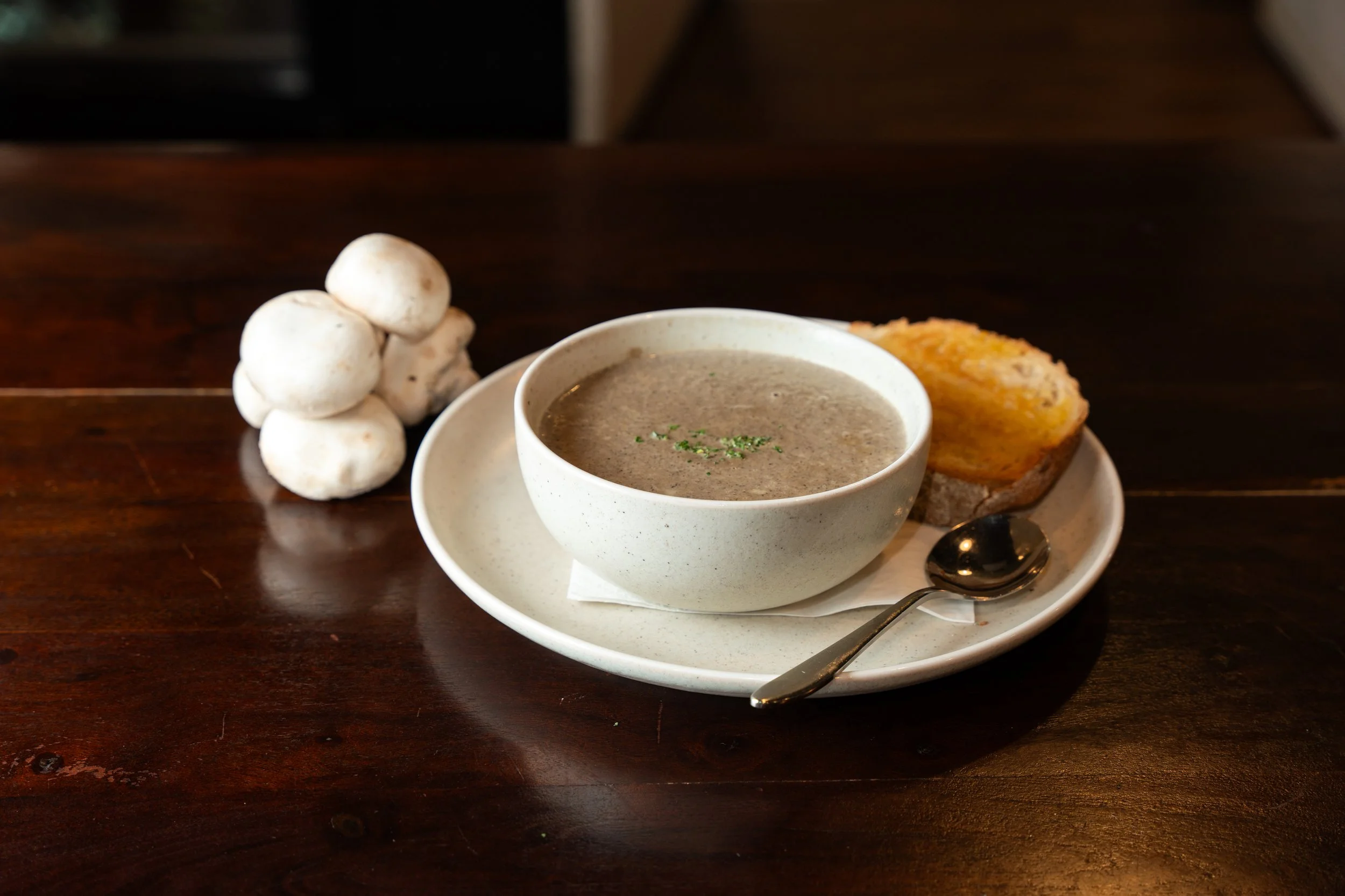 A bowl of mushroom soup on a white plate with a spoon, a slice of bread topped with cheese, a bunch of mushrooms, on a dark wooden table.
