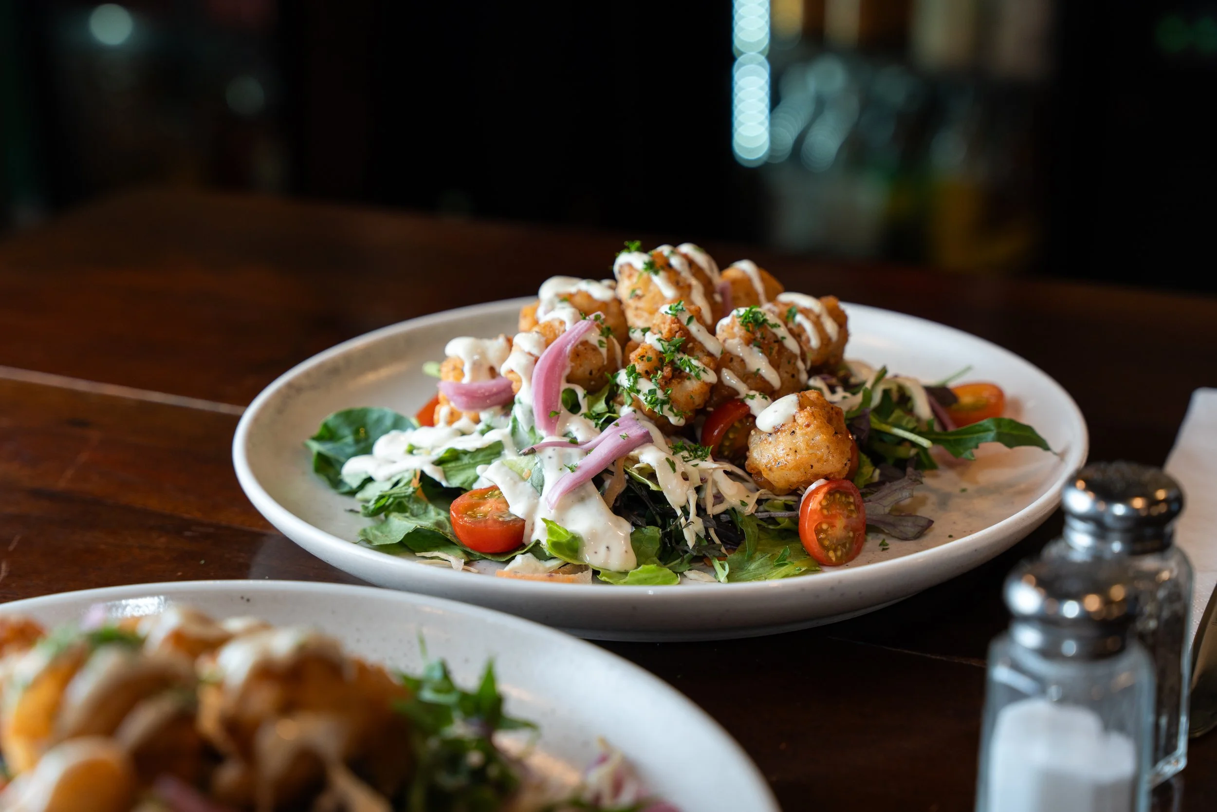 Plate of fried chicken pieces with creamy white dressing, served on a bed of mixed greens, cherry tomatoes, and sliced red onions in a restaurant setting.