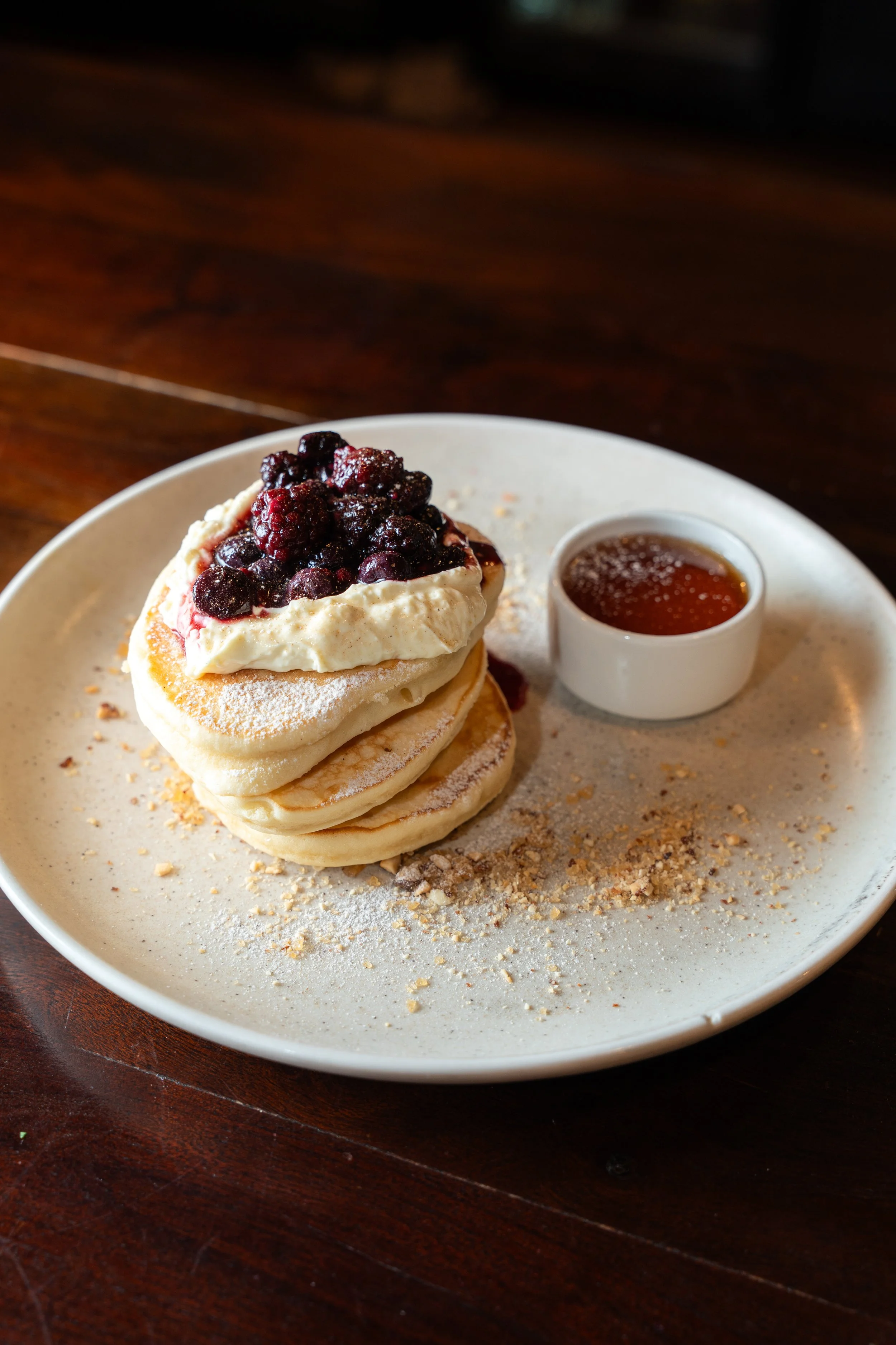 Stack of three pancakes topped with whipped cream and mixed berries, served with honey or syrup on a white plate.