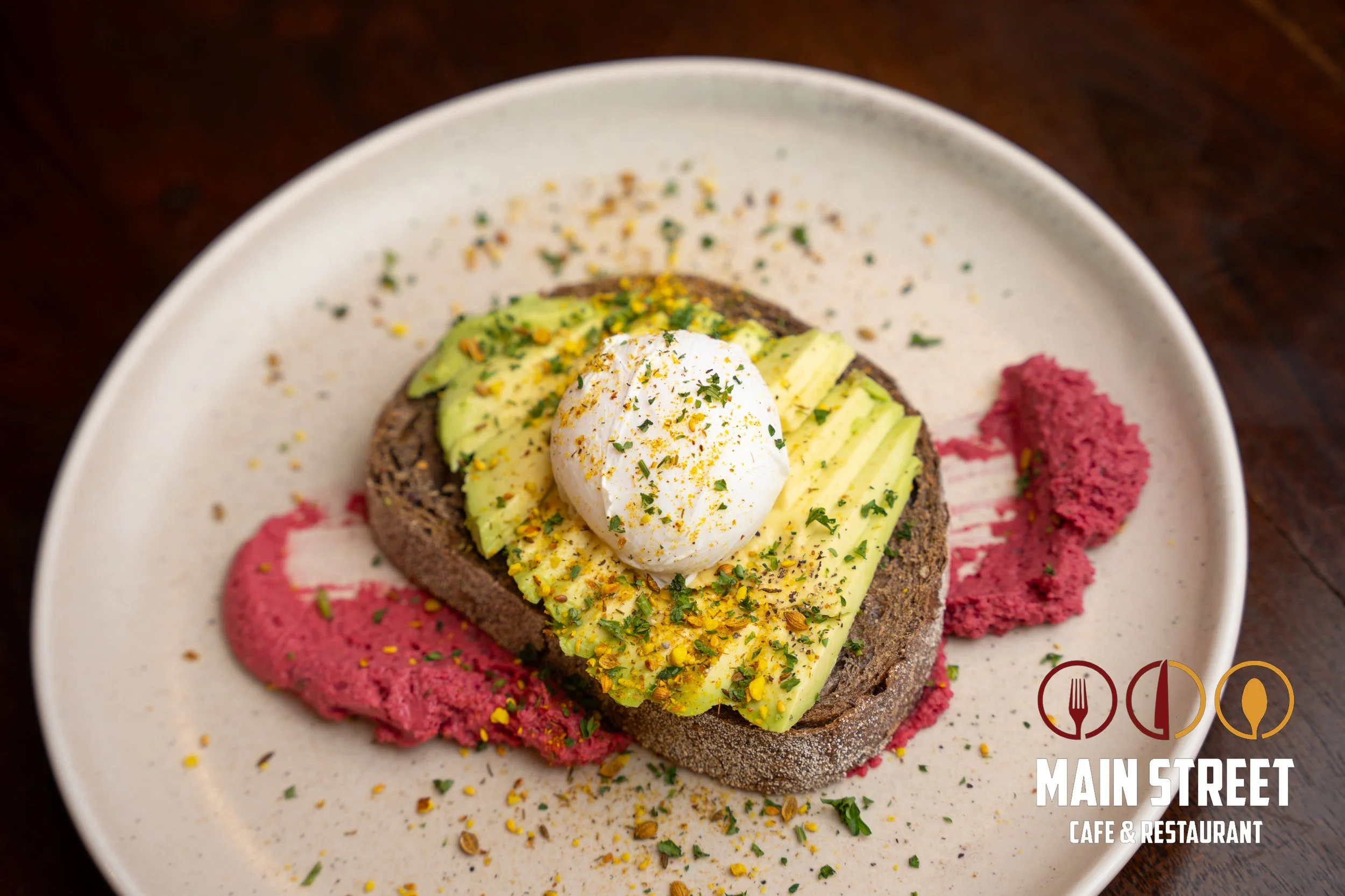 Sourdough toast topped with avocado slices, poached egg, and sprinkled herbs, served with pink beet hummus, on a white plate from Main Street Cafe & Restaurant.
