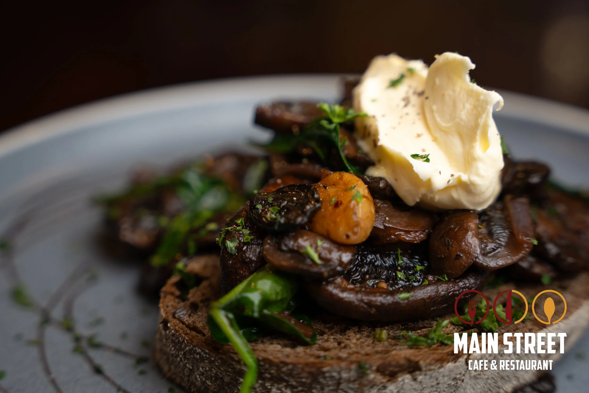 Close-up of a toasted bread slice topped with sautéed mushrooms, herbs, a dollop of butter or cheese, garnished with parsley, on a plate at Main Street Café & Restaurant.
