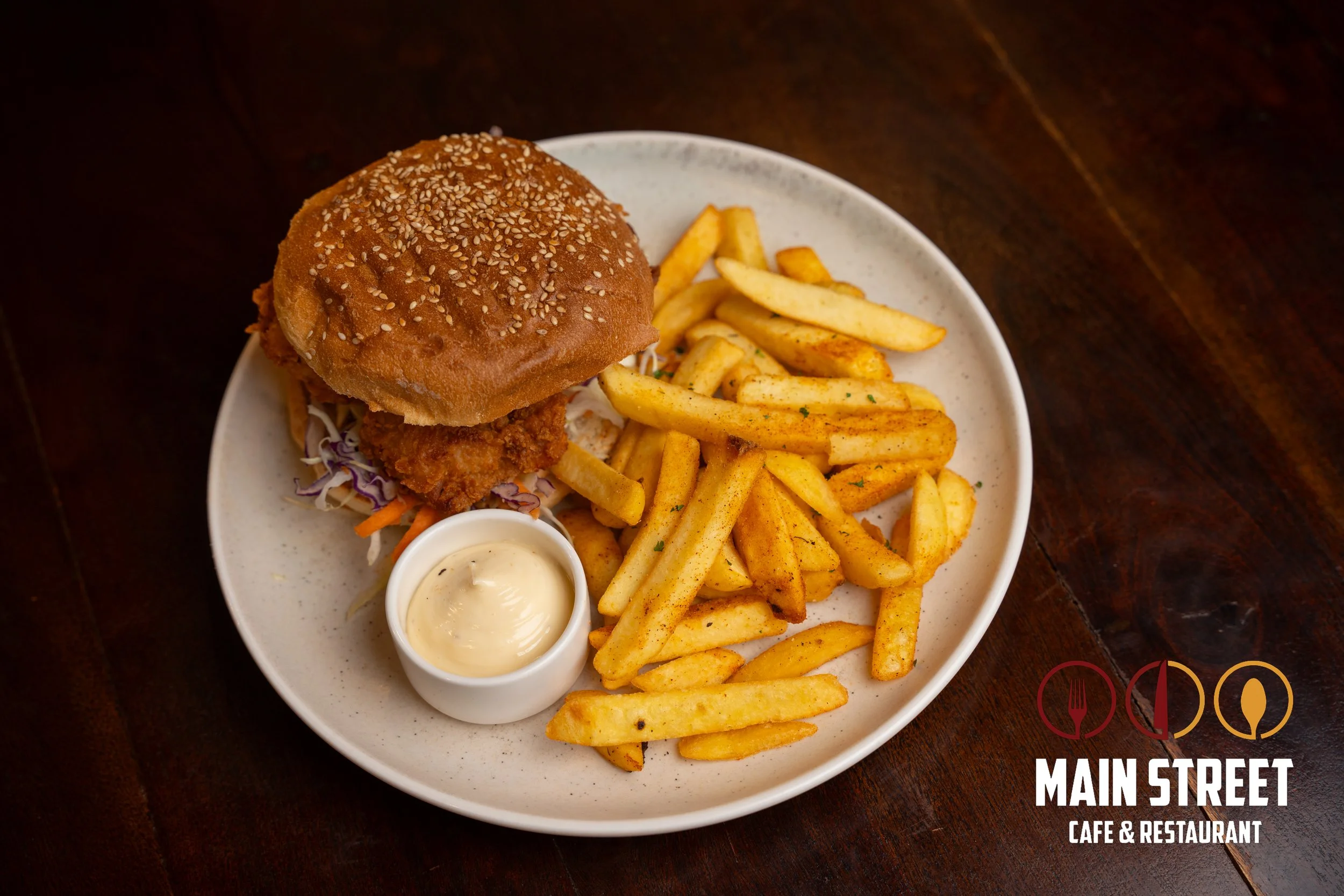 Fried chicken sandwich with lettuce and sauce in a sesame seed bun, served with seasoned French fries and a side of dipping sauce on a white plate.