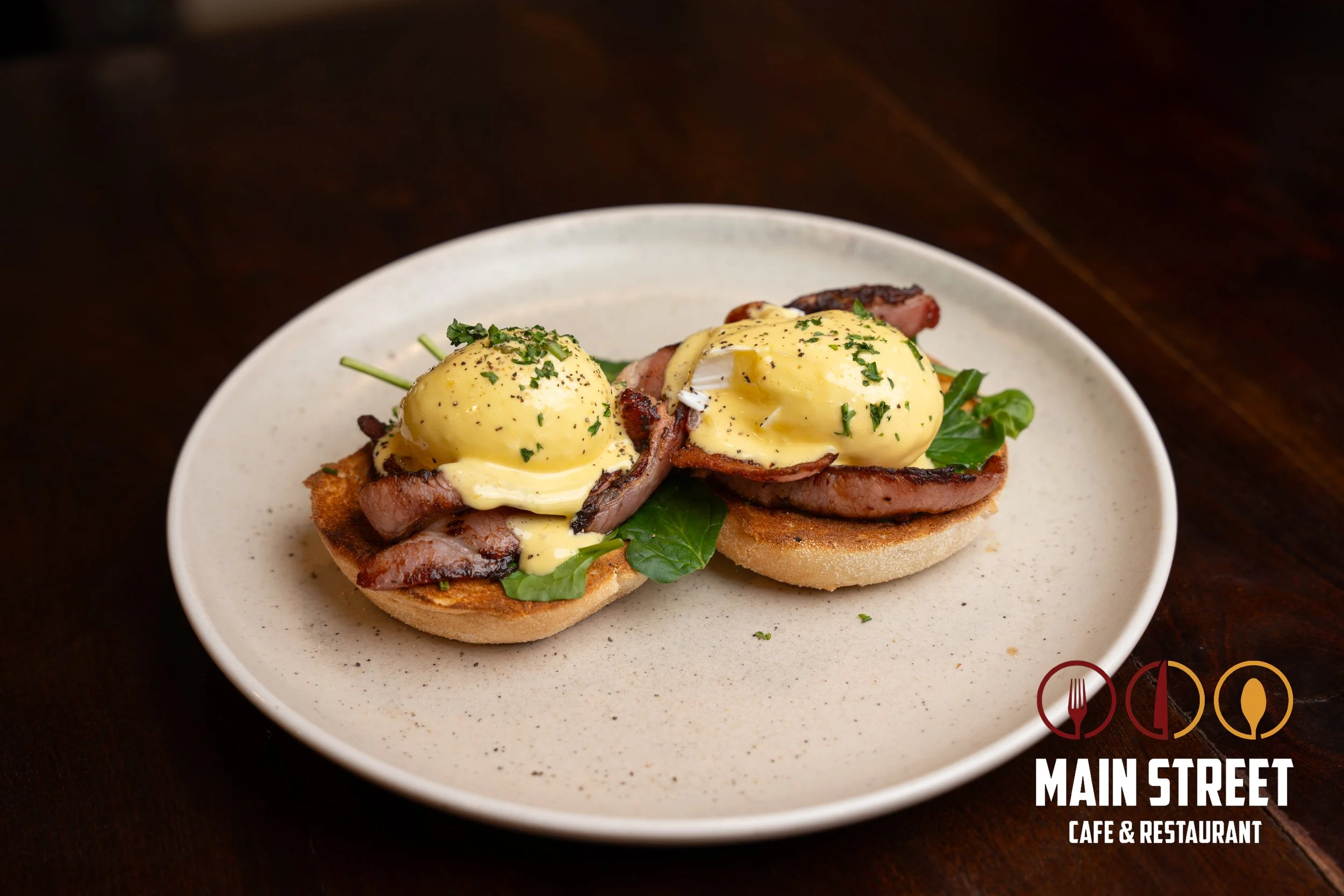 Two eggs Benedict with bacon, greens, and hollandaise sauce on toasted bread on a white plate, with a logo for Main Street Cafe & Restaurant in the bottom right corner.