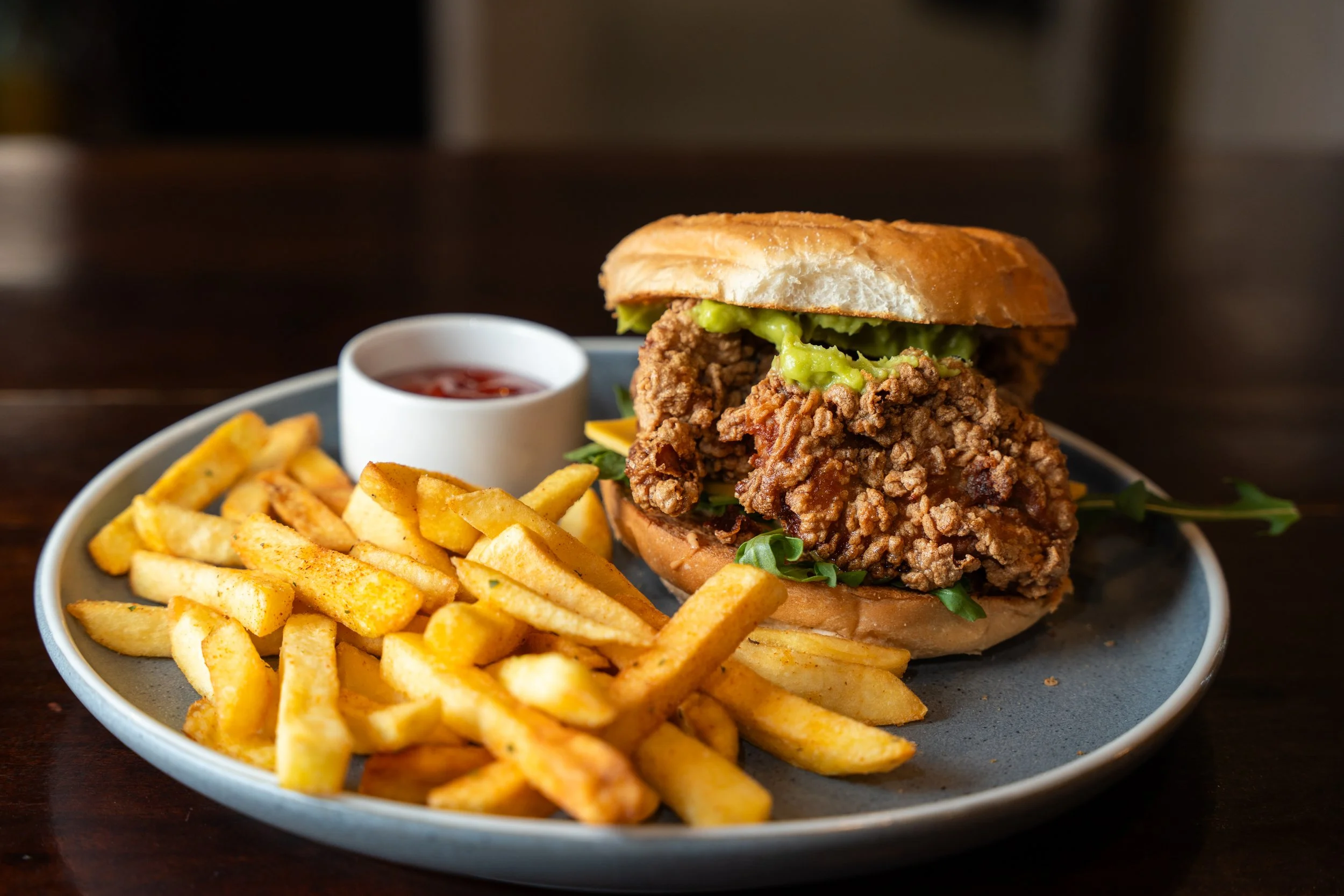 A serving of fried chicken sandwich with lettuce, pickles, and bun on a gray plate, accompanied by French fries and a small cup of ketchup.