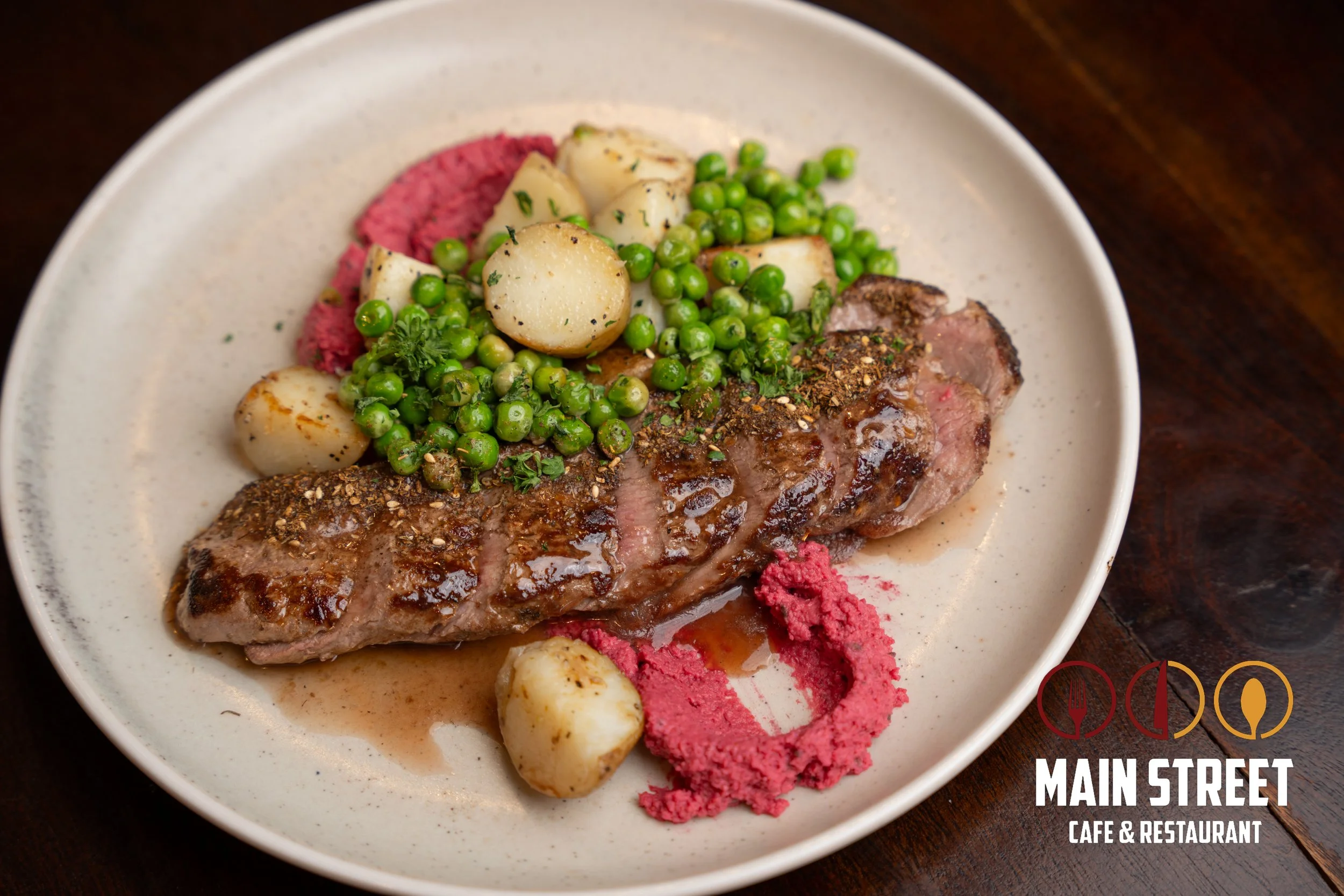 Plate with cooked steak, green peas, roasted potatoes, and colorful purees on a white dish, with a Main Street Cafe & Restaurant.