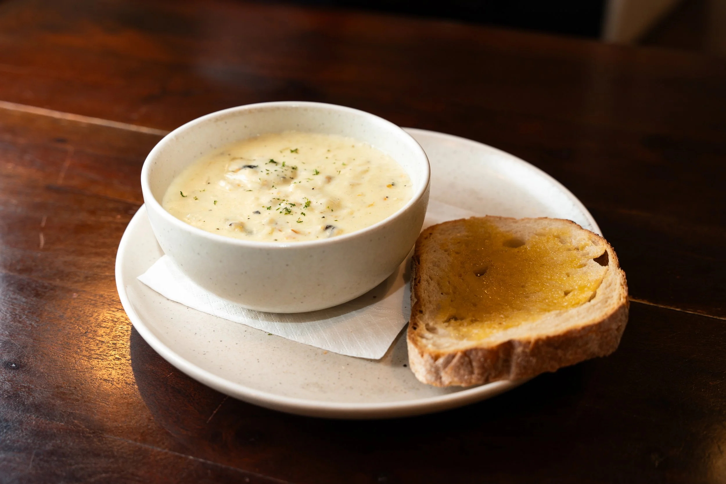Creamy soup with herbs in a white bowl, served with a slice of toasted bread with butter on a white plate on a wooden table.