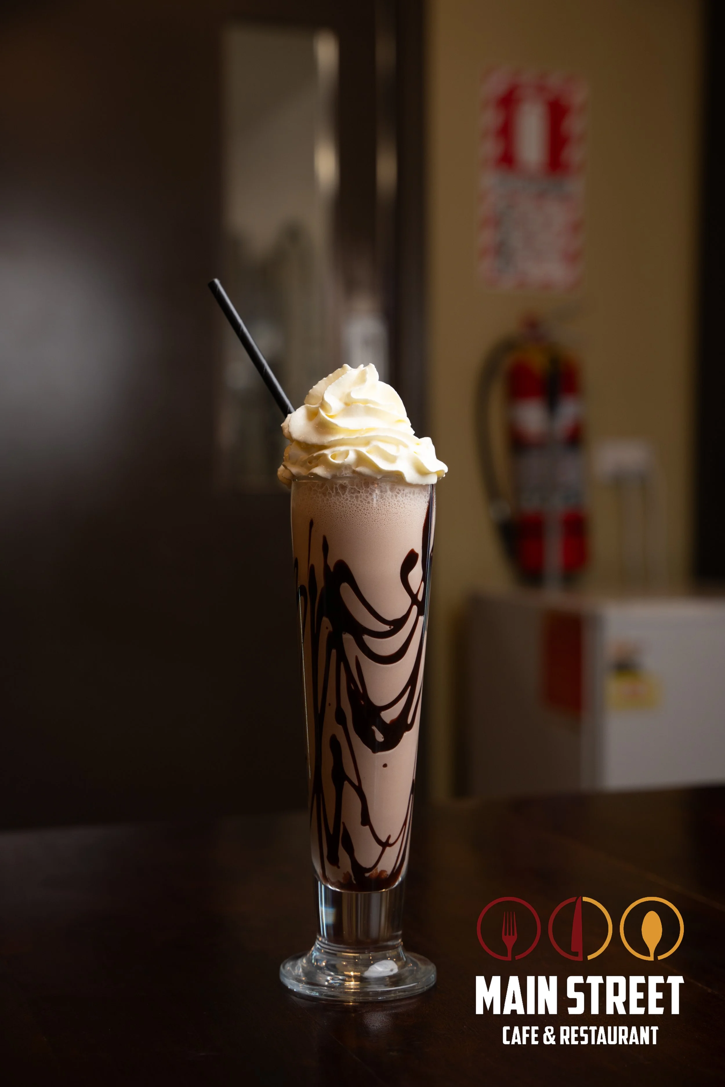 A tall glass of chocolate milkshake topped with whipped cream and chocolate syrup swirl, with a black straw. The glass is placed on a wooden table in a café or restaurant, with a logo of Main Street Café & Restaurant in the bottom right corner.