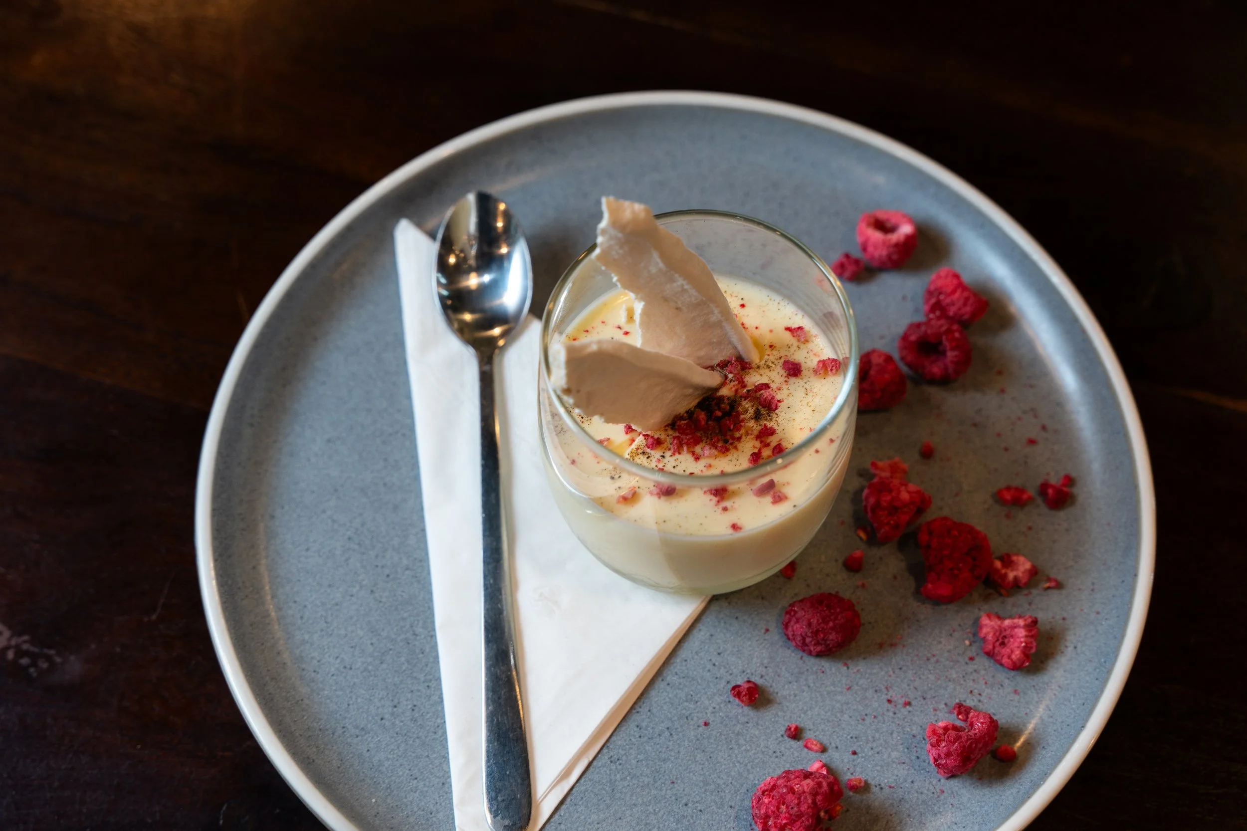 Dessert in a glass topped with white chocolate shavings and pink crumbles, served on a gray plate with scattered raspberries, a spoon, and a folded napkin.