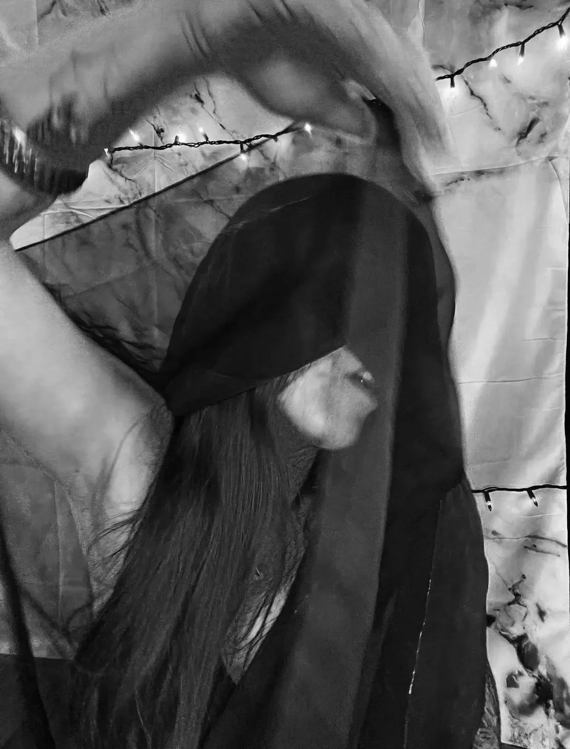 A woman with long hair wearing a dark hooded jacket, covered in a black mesh veil, supporting her head with her arms, in a setting decorated with string lights.