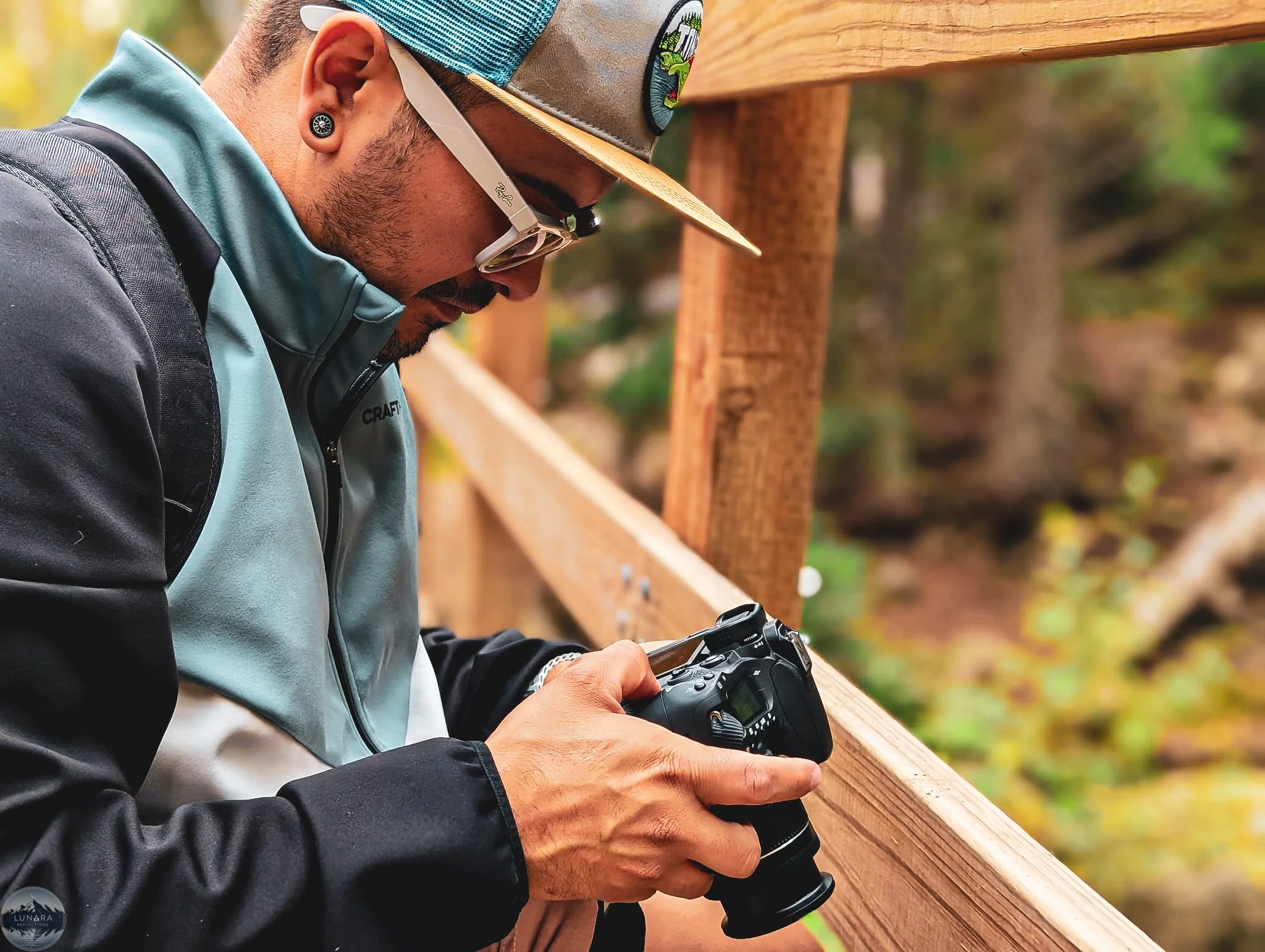 Man with a camera taking photos outdoors, wearing sunglasses, a cap, and a jacket, leaning on a wooden railing with trees in the background.