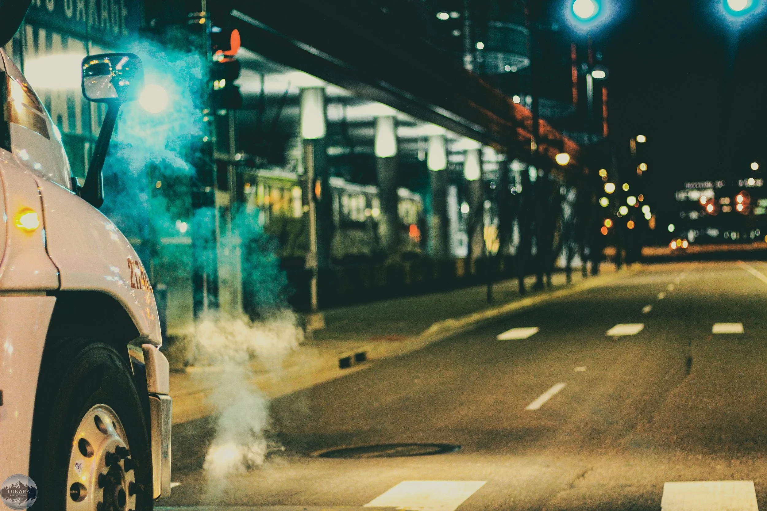 A city street at night with a steam-cleaned bus lane, visible reflecting city lights, and a steam cleaning truck emitting white vapor, while the bus stop features illuminated signage and modern glass architecture.
