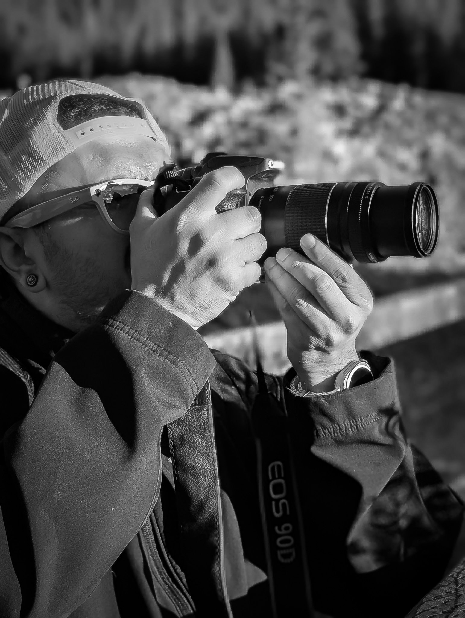A person wearing a cap and glasses taking a photo with a camera outdoors in black and white.