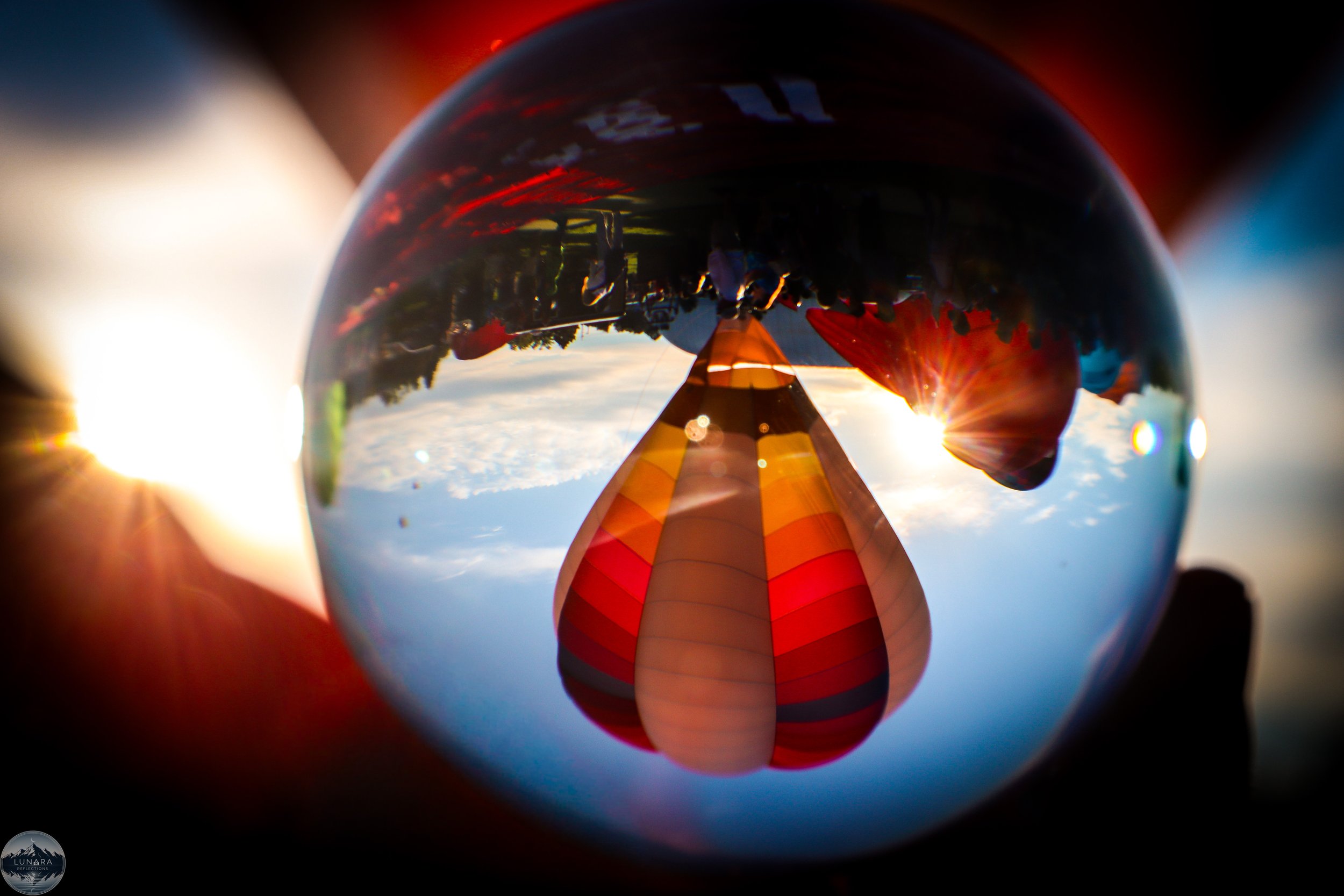 A hot air balloon with red, orange, and yellow stripes reflected in a glass sphere, with the sky and clouds visible in the background.